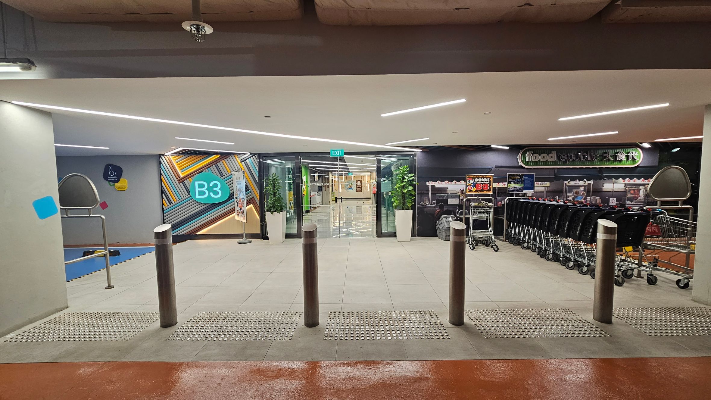On the left, a wall with a colourful geometric design and a sign marked "B3". In the centre, glass doors with an "EXIT" sign above, flanked by potted plants. On the right, a row of shopping trolleys and a sign for "food republic". The floor is tiled with tactile paving near the entrance, and the ceiling has modern strip lighting. On the left, a wall with a colourful geometric design and a sign marked "B3". In the centre, glass doors with an "EXIT" sign above, flanked by potted plants. On the right, a row of shopping trolleys and a sign for "food republic". The floor is tiled with tactile paving near the entrance, and the ceiling has modern strip lighting.