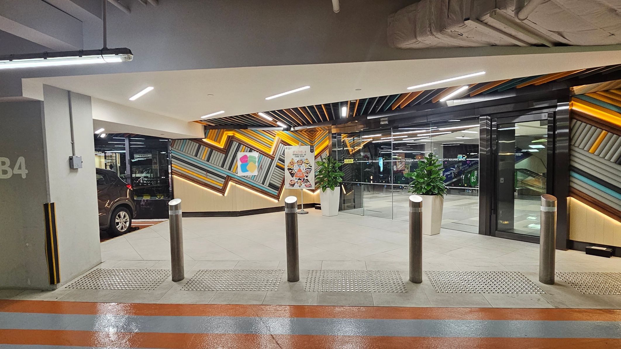 A modern car park entrance with a geometric, colourful wall design featuring diagonal lines in shades of orange, blue, and grey. On the left, a black car is partially visible next to a grey pillar marked "B4". In the centre, there are three metallic bollards and a sign with images of food. Two potted plants are placed near the glass doors on the right, leading to an interior space. The ceiling has exposed pipes and bright lighting. The floor is tiled with a textured strip near the bollards. A modern car park entrance with a geometric, colourful wall design featuring diagonal lines in shades of orange, blue, and grey. On the left, a black car is partially visible next to a grey pillar marked "B4". In the centre, there are three metallic bollards and a sign with images of food. Two potted plants are placed near the glass doors on the right, leading to an interior space. The ceiling has exposed pipes and bright lighting. The floor is tiled with a textured strip near the bollards.