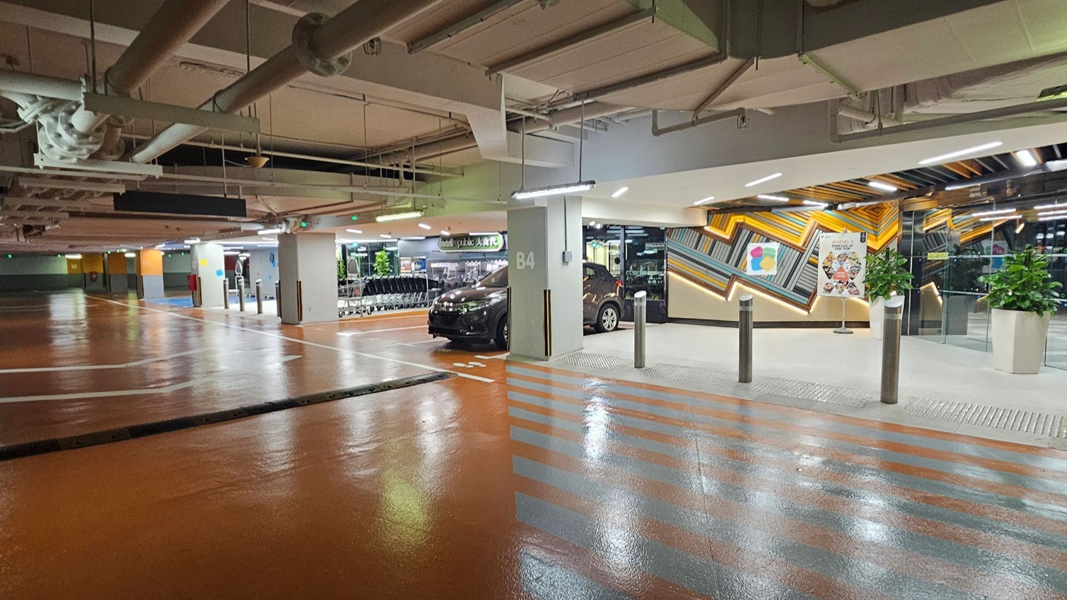 A brightly lit underground car park with a shiny, polished floor. On the left, there are empty parking spaces and a row of shopping trolleys. In the centre, a black car is parked next to a pillar marked "B4". On the right, a colourful geometric wall design with a poster and two large potted plants. The ceiling is exposed with visible pipes and lighting fixtures. A brightly lit underground car park with a shiny, polished floor. On the left, there are empty parking spaces and a row of shopping trolleys. In the centre, a black car is parked next to a pillar marked "B4". On the right, a colourful geometric wall design with a poster and two large potted plants. The ceiling is exposed with visible pipes and lighting fixtures.