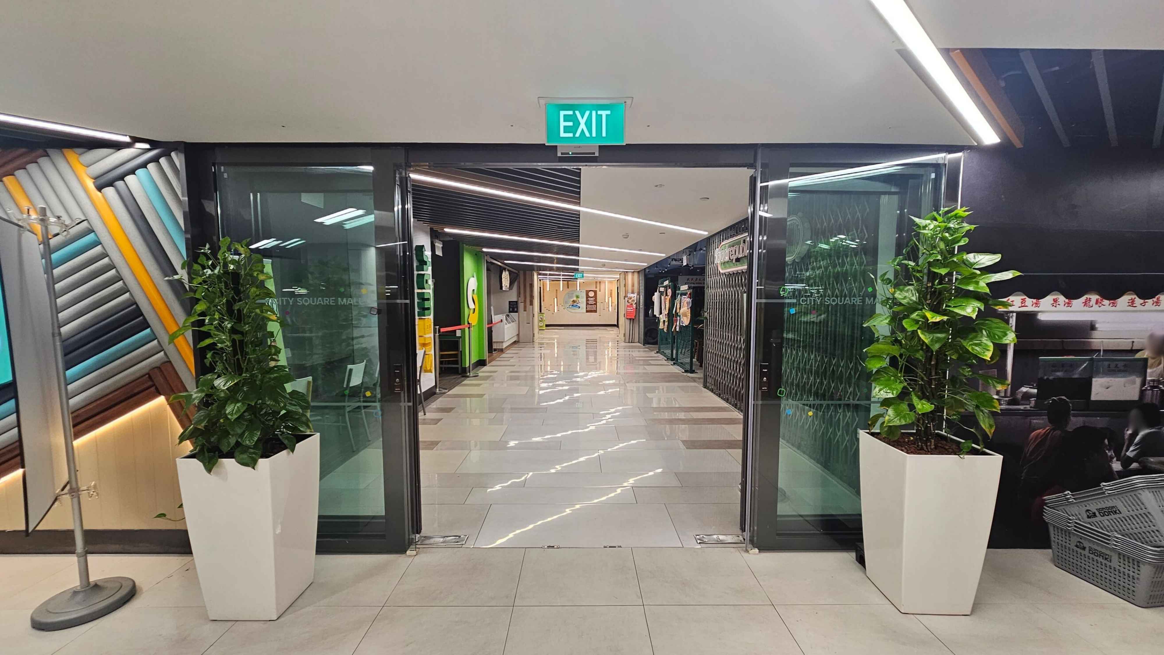 Glass doors with an "EXIT" sign above them, flanked by tall green plants in white pots. Beyond the doors, a corridor with a shiny tiled floor leads to various shops. On the left, colourful diagonal wall patterns, and on the right, a dark wall with a partially visible food stall. The ceiling features linear lighting. Glass doors with an "EXIT" sign above them, flanked by tall green plants in white pots. Beyond the doors, a corridor with a shiny tiled floor leads to various shops. On the left, colourful diagonal wall patterns, and on the right, a dark wall with a partially visible food stall. The ceiling features linear lighting.