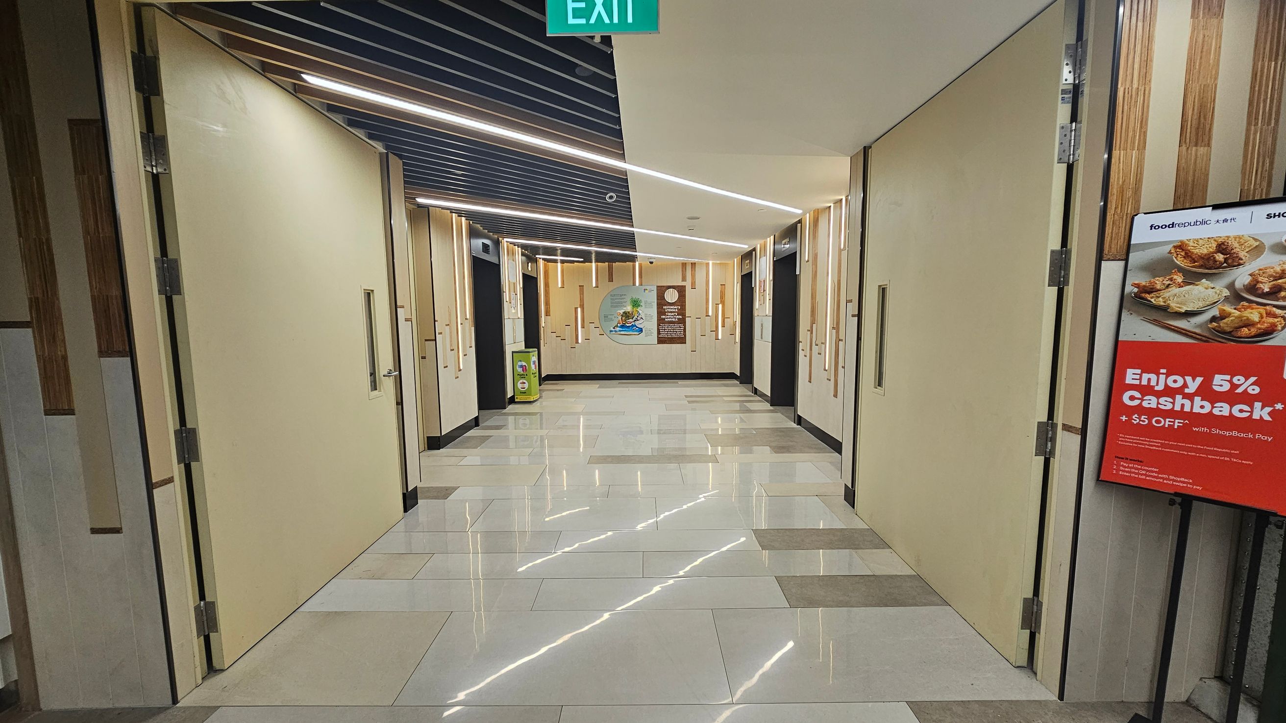 A corridor with beige walls and large doors on both sides. The floor is tiled with a glossy finish, reflecting the ceiling lights. The ceiling has a modern design with linear lights and dark panels. On the right, there's a poster advertising a 5% cashback offer with images of food. At the end of the corridor, there's a mural and a small green bin. An exit sign is visible at the top. A corridor with beige walls and large doors on both sides. The floor is tiled with a glossy finish, reflecting the ceiling lights. The ceiling has a modern design with linear lights and dark panels. On the right, there's a poster advertising a 5% cashback offer with images of food. At the end of the corridor, there's a mural and a small green bin. An exit sign is visible at the top.