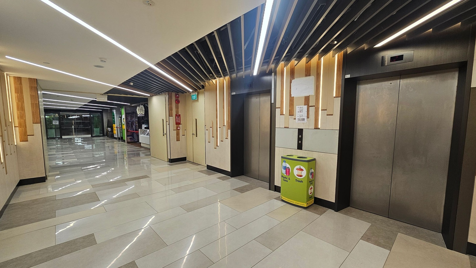 A modern indoor hallway with a polished tiled floor and a ceiling featuring linear lights. On the right, there are two lift doors with a green recycling bin nearby. The left side shows a corridor leading to glass doors, with a fire extinguisher mounted on the wall. The walls have a light wood and beige design, and the ceiling has black slats with integrated lighting. A modern indoor hallway with a polished tiled floor and a ceiling featuring linear lights. On the right, there are two lift doors with a green recycling bin nearby. The left side shows a corridor leading to glass doors, with a fire extinguisher mounted on the wall. The walls have a light wood and beige design, and the ceiling has black slats with integrated lighting.