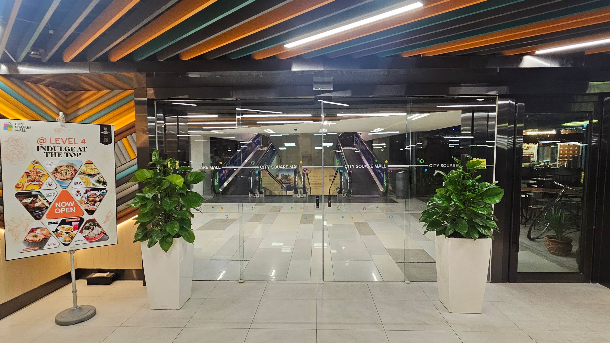 Glass entrance doors to City Square Mall with escalators visible inside. On the left, a colourful sign advertises dining options on Level 4, featuring various dishes and a "Now Open" message. Two large potted plants flank the entrance. The ceiling has a pattern of orange, green, and black slats, and the floor is tiled in light colours. Glass entrance doors to City Square Mall with escalators visible inside. On the left, a colourful sign advertises dining options on Level 4, featuring various dishes and a "Now Open" message. Two large potted plants flank the entrance. The ceiling has a pattern of orange, green, and black slats, and the floor is tiled in light colours.