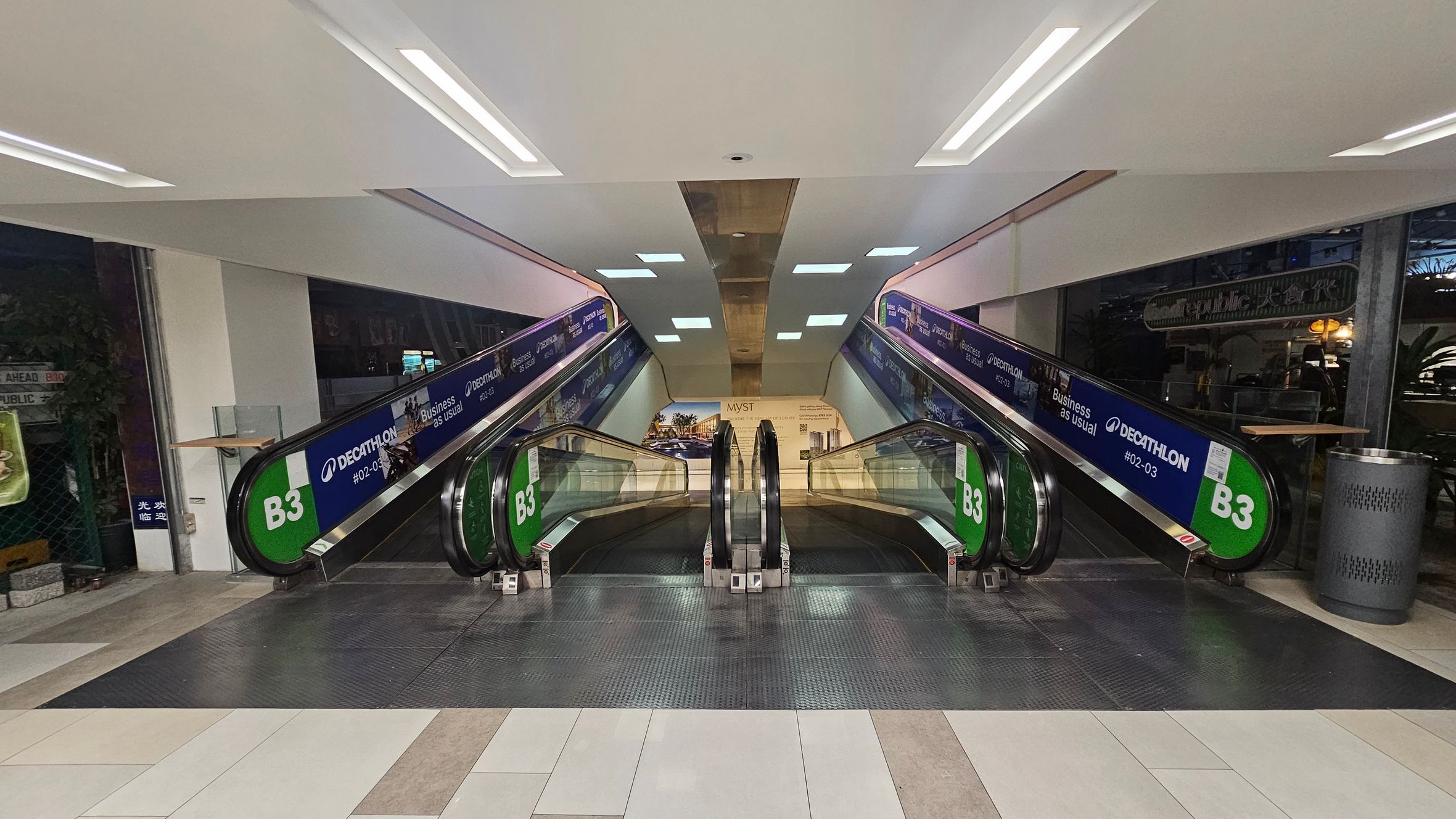 Two parallel escalators in a shopping centre, with green signs marked "B3" on the sides. The left escalator is for descending, and the right is for ascending. Advertisements for a store are visible along the sides. The floor is tiled in a mix of light and dark shades, and the ceiling is white with recessed lighting. A trash bin is on the right side near the escalator. In the background, there is a large poster with images and text. Two parallel escalators in a shopping centre, with green signs marked "B3" on the sides. The left escalator is for descending, and the right is for ascending. Advertisements for a store are visible along the sides. The floor is tiled in a mix of light and dark shades, and the ceiling is white with recessed lighting. A trash bin is on the right side near the escalator. In the background, there is a large poster with images and text.