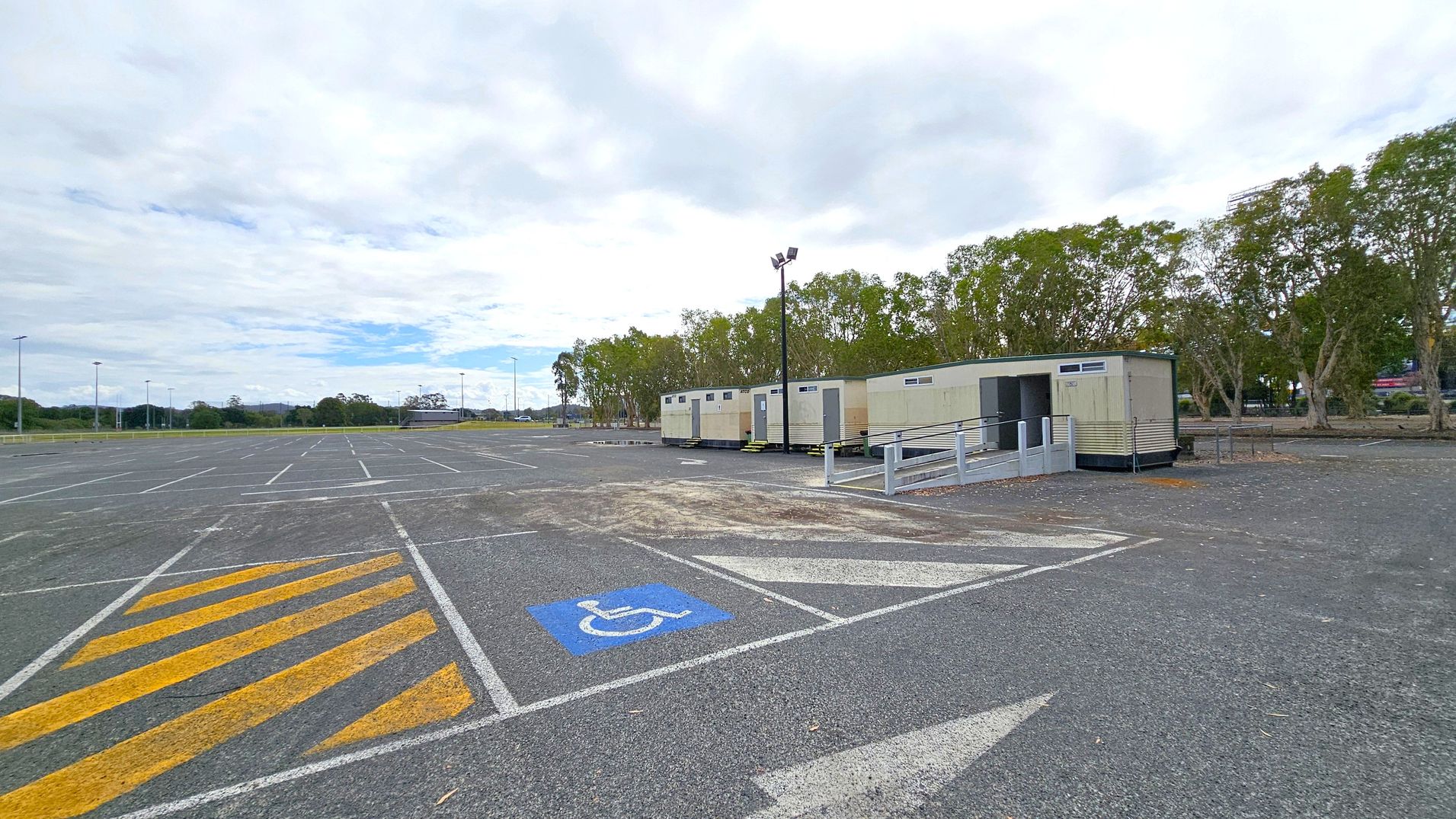 A large, mostly empty car park with marked parking spaces, including a blue disabled parking spot in the foreground. Yellow diagonal lines are painted on the ground nearby. On the right, several portable buildings are lined up, with a ramp leading to one of them. Trees are visible in the background, and a few streetlights are scattered throughout the area. The sky is overcast.