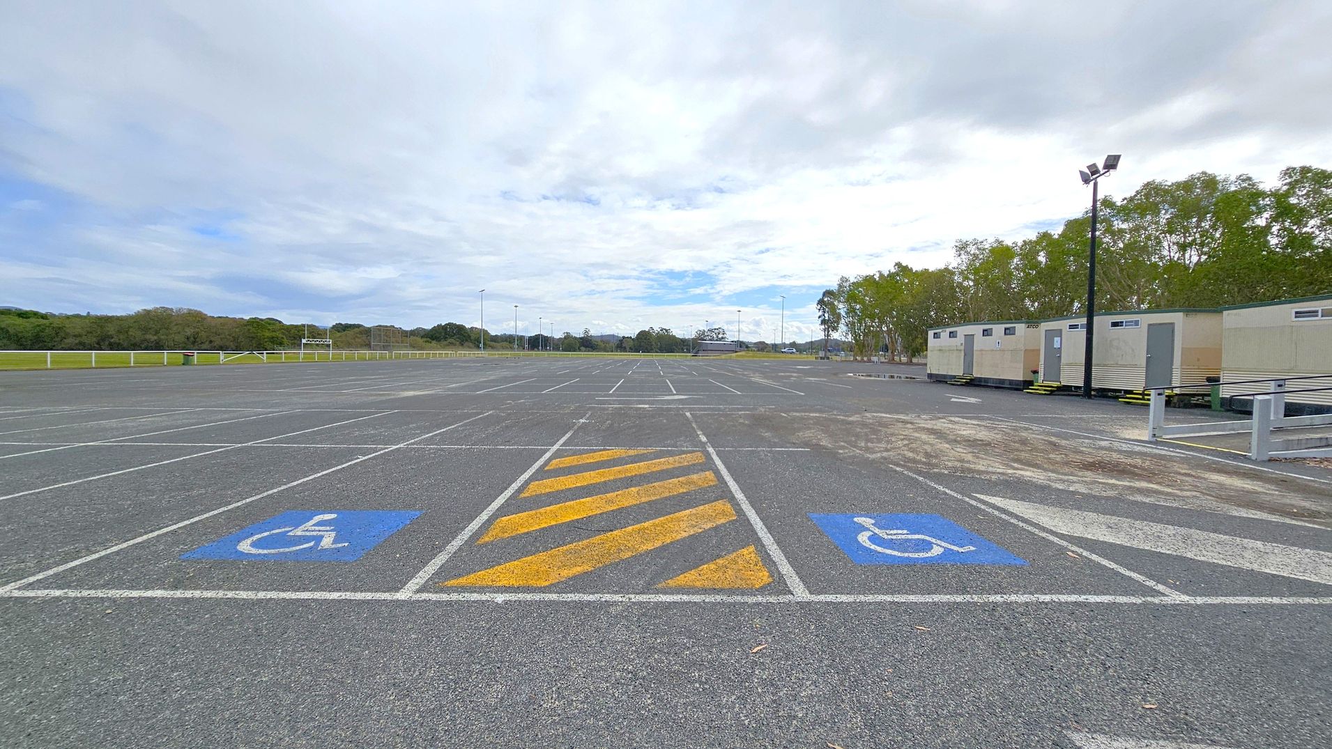 A large, empty car park with marked parking spaces, including two blue disabled parking spaces and a yellow-striped area in the foreground. On the right, there are several portable buildings lined up, with a row of trees behind them. A tall lamp post is visible near the buildings. The background features more trees and open fields, with a few distant structures and light poles. The sky is overcast.