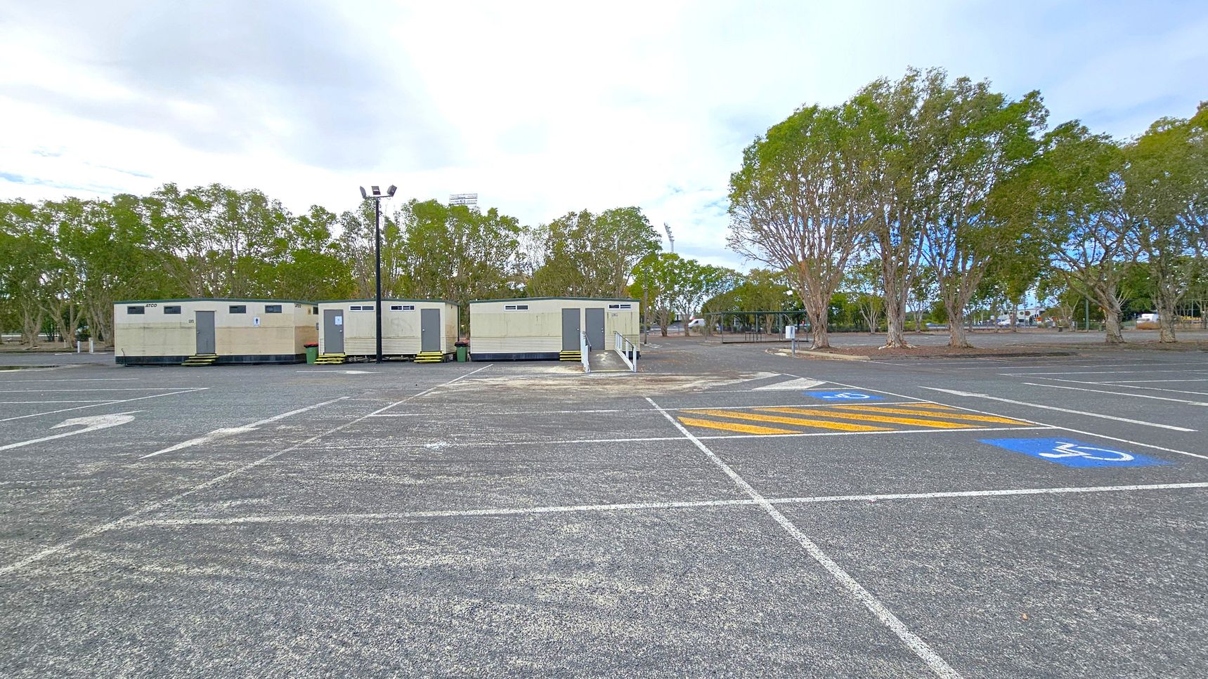 A large, empty car park with marked spaces, including a disabled parking area with blue and yellow markings in the foreground. In the middle, there are three portable buildings with steps leading up to their doors. A tall light pole stands nearby. In the background, there are several trees with green foliage lining the edge of the car park. The ground is asphalt, and the area appears spacious and open.