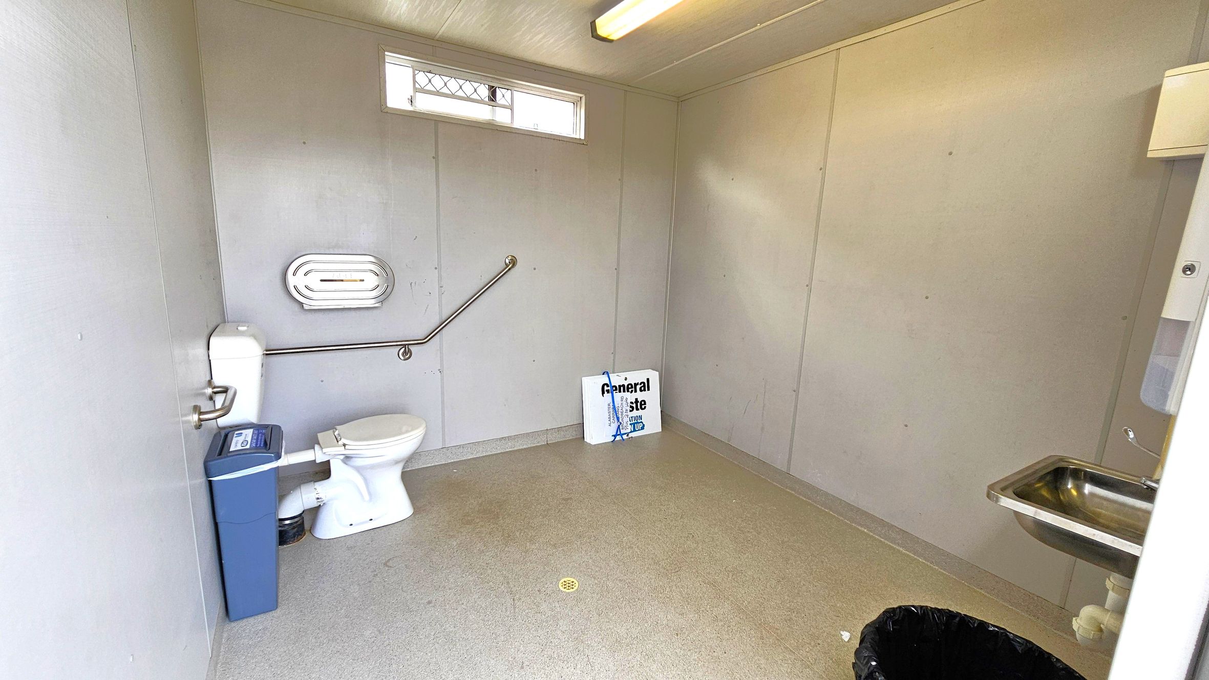 A spacious public toilet with light-coloured walls and a non-slip floor. On the left, there's a white toilet with a grab rail and a blue sanitary bin beside it. A small window with a mesh cover is above the toilet. In the centre, a sign reads "General Waste." On the right, a stainless steel sink with a soap dispenser is mounted on the wall. A black rubbish bin is on the floor near the sink. The ceiling has a fluorescent light fixture.
