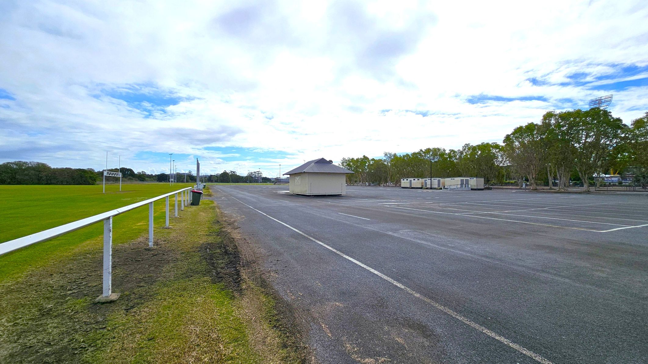 On the left, there's a grassy field with a white railing and a rubbish bin. In the middle, a small, light-coloured building sits on a large, empty paved area. On the right, several portable buildings are lined up near a row of trees. The ground is a mix of grass and asphalt.
