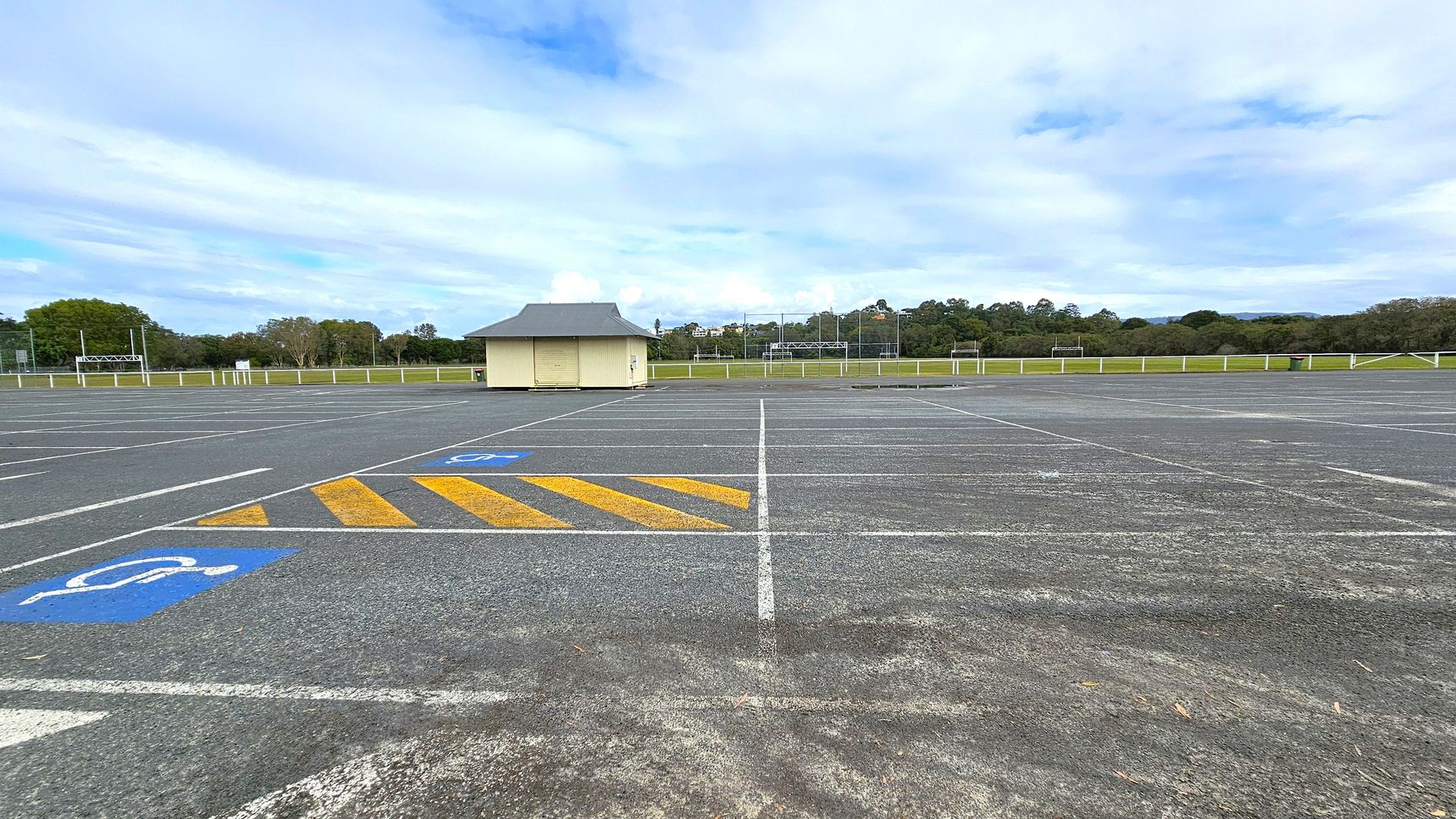 An empty car park with marked parking spaces, including a blue disabled parking symbol on the left. Yellow diagonal lines are painted near the symbol. In the centre, a small, light-coloured building with a dark roof stands alone. The background features a grassy field bordered by trees and a fence. The sky is overcast with patches of blue.