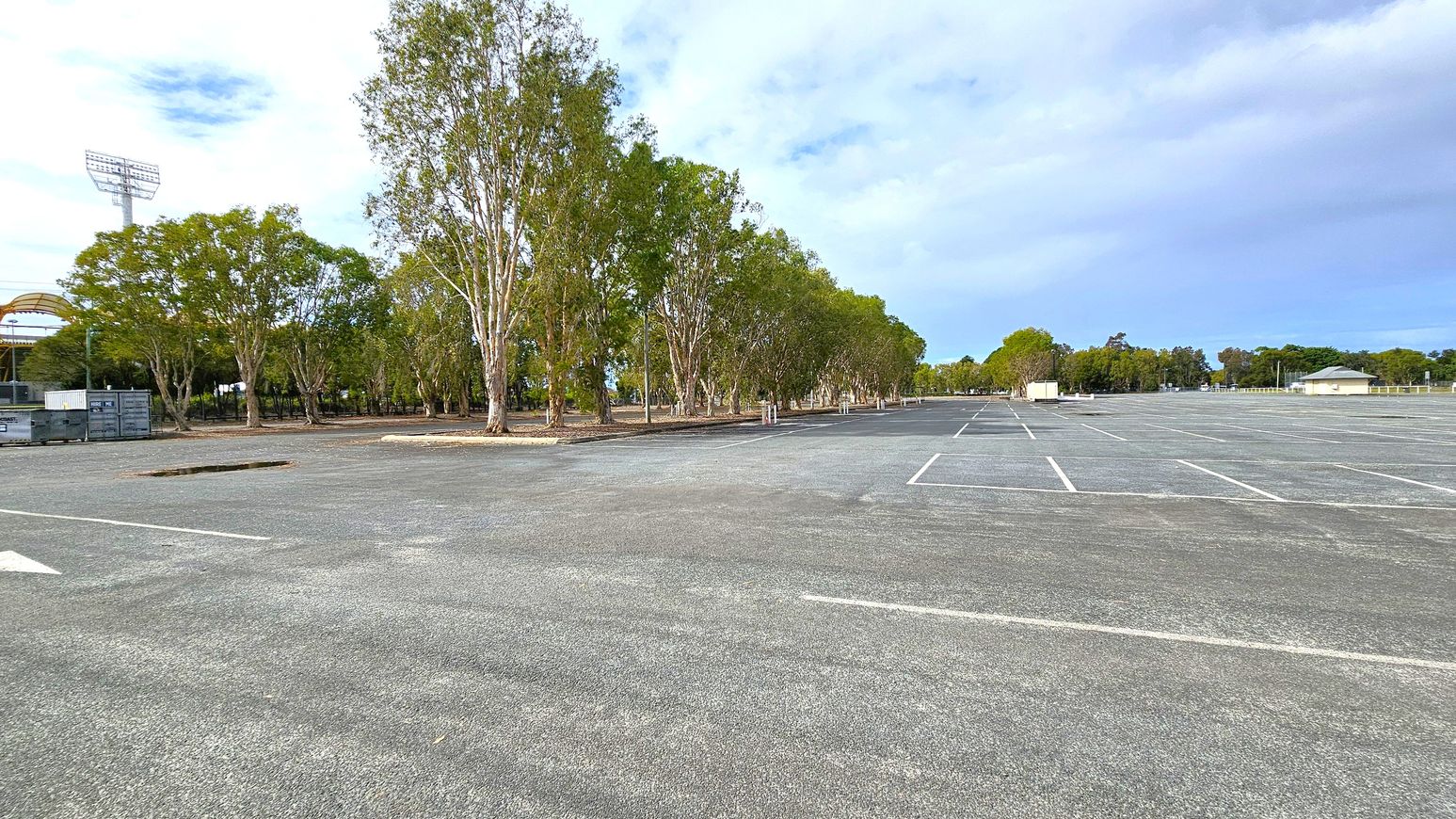 A large, empty car park with marked parking spaces stretches across the foreground. On the left, there are several tall trees and a few shipping containers. A stadium light tower is visible in the distance. The right side features more trees and a small building with a green roof. The background is filled with additional trees and open space. The sky is overcast.