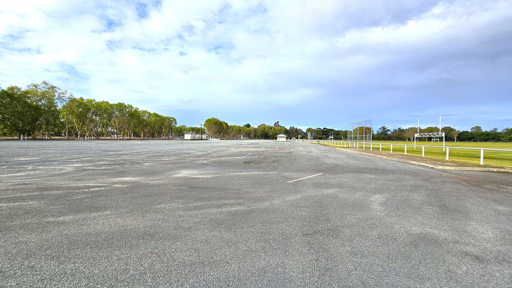 A large, empty car park with a grey asphalt surface stretches across the foreground. On the left, a row of trees lines the edge, providing a natural border. In the middle, there are small structures, possibly sheds or kiosks. On the right, a grassy field is enclosed by a white fence, with a scoreboard and goalposts visible. The background features more trees, creating a green backdrop. The sky is overcast, casting a soft light over the scene.