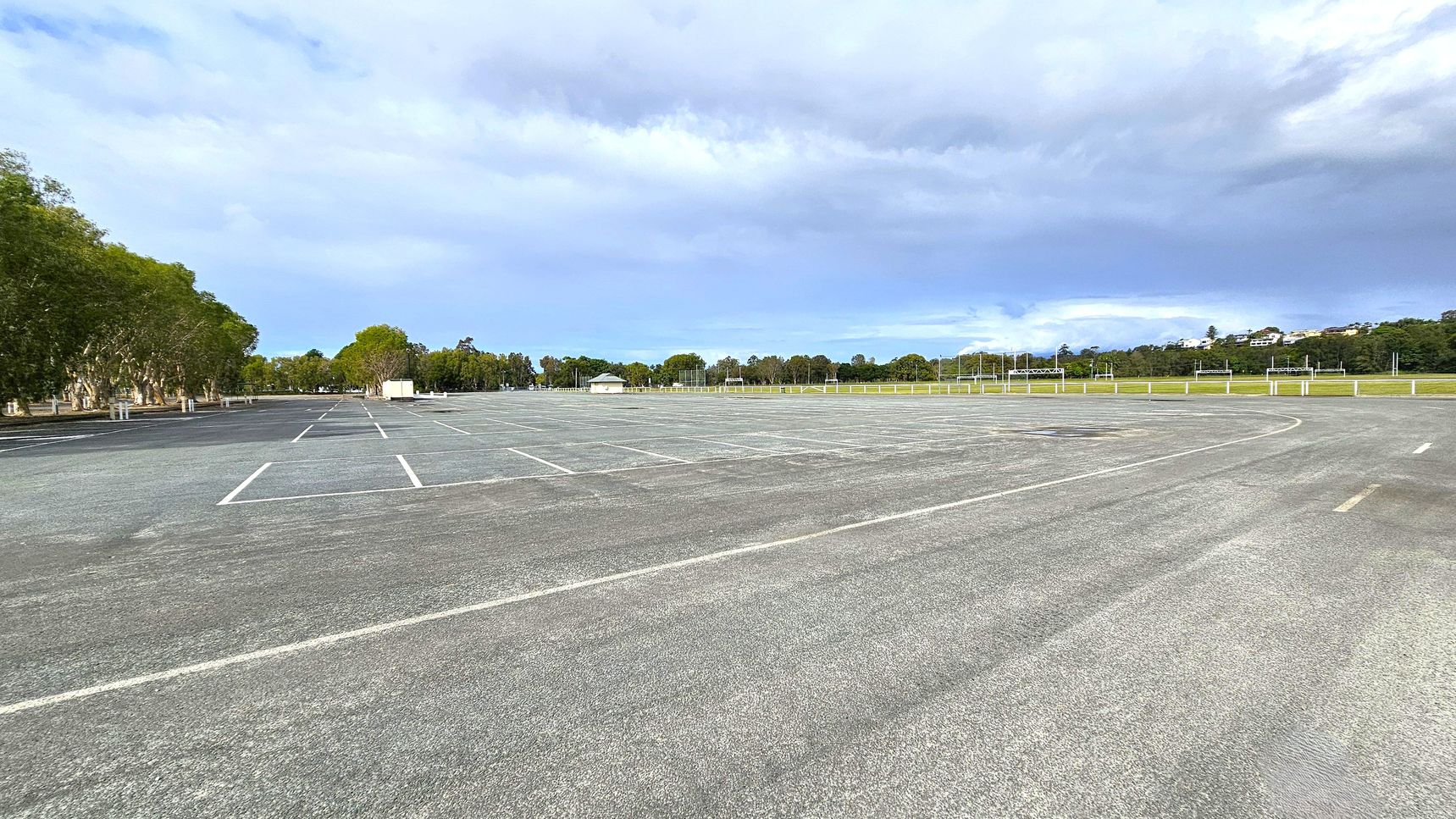 An expansive, empty car park with marked parking spaces on an asphalt surface. On the left, there are trees lining the edge of the car park. In the background, a small white building is visible, surrounded by more trees and a grassy area. The right side features a grassy field with a fence and some distant buildings on a hill. The sky is overcast, casting a soft light over the scene.