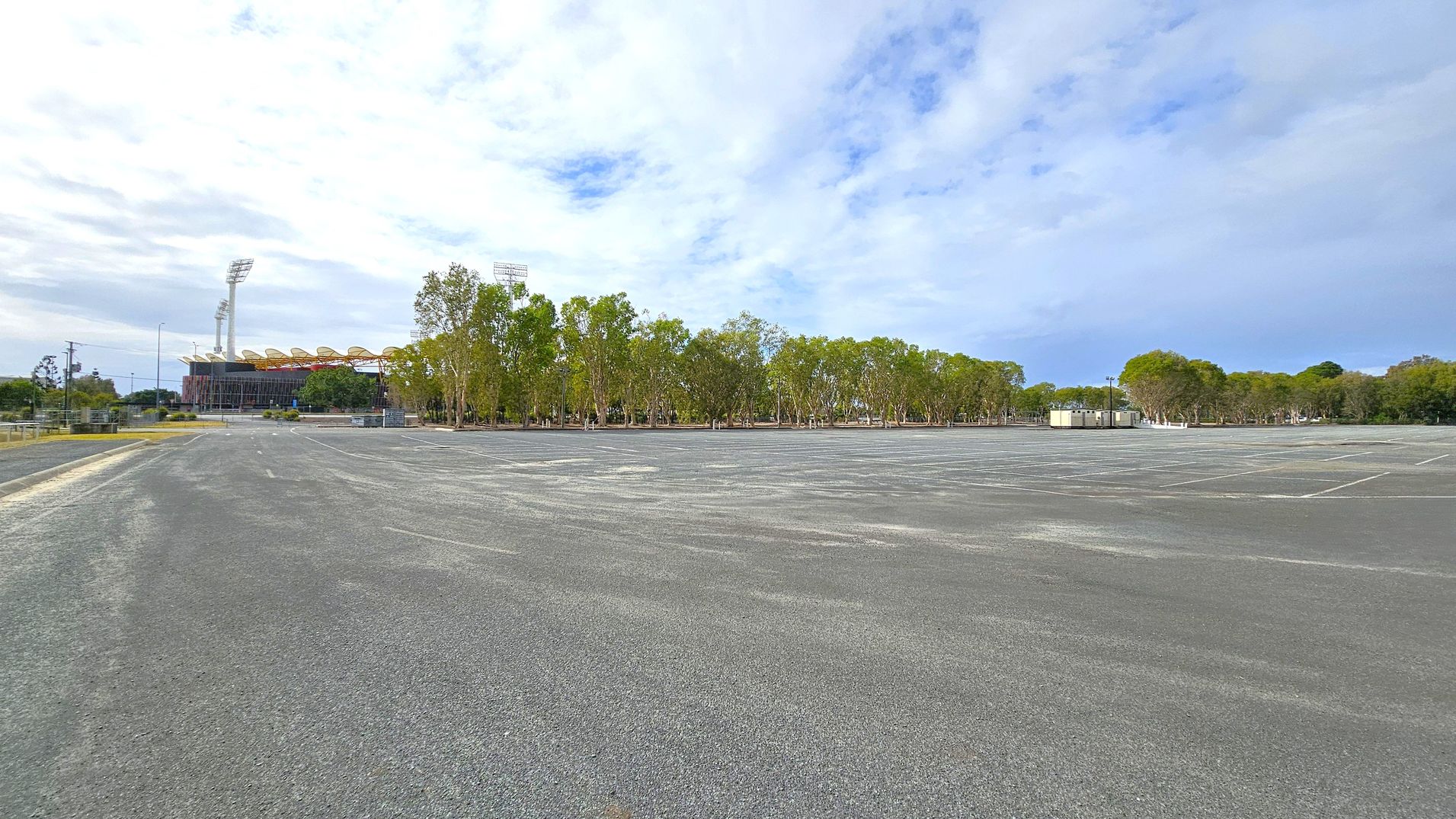 A large, empty car park with a grey asphalt surface stretches across the foreground. In the background, there are rows of tall trees with green foliage. To the left, a stadium with tall floodlights and a distinctive roof structure is visible. On the right, there are a few small, light-coloured portable buildings. The sky is mostly clear with some clouds.