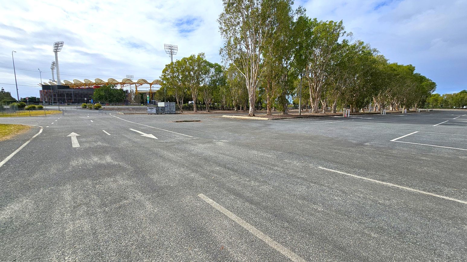 A large, empty car park with marked parking spaces and directional arrows on the asphalt. On the left, a stadium with tall floodlights and a distinctive roof structure is visible. To the right, a row of tall trees lines the edge of the car park, providing a natural border. The ground is paved, and the area appears spacious and open.