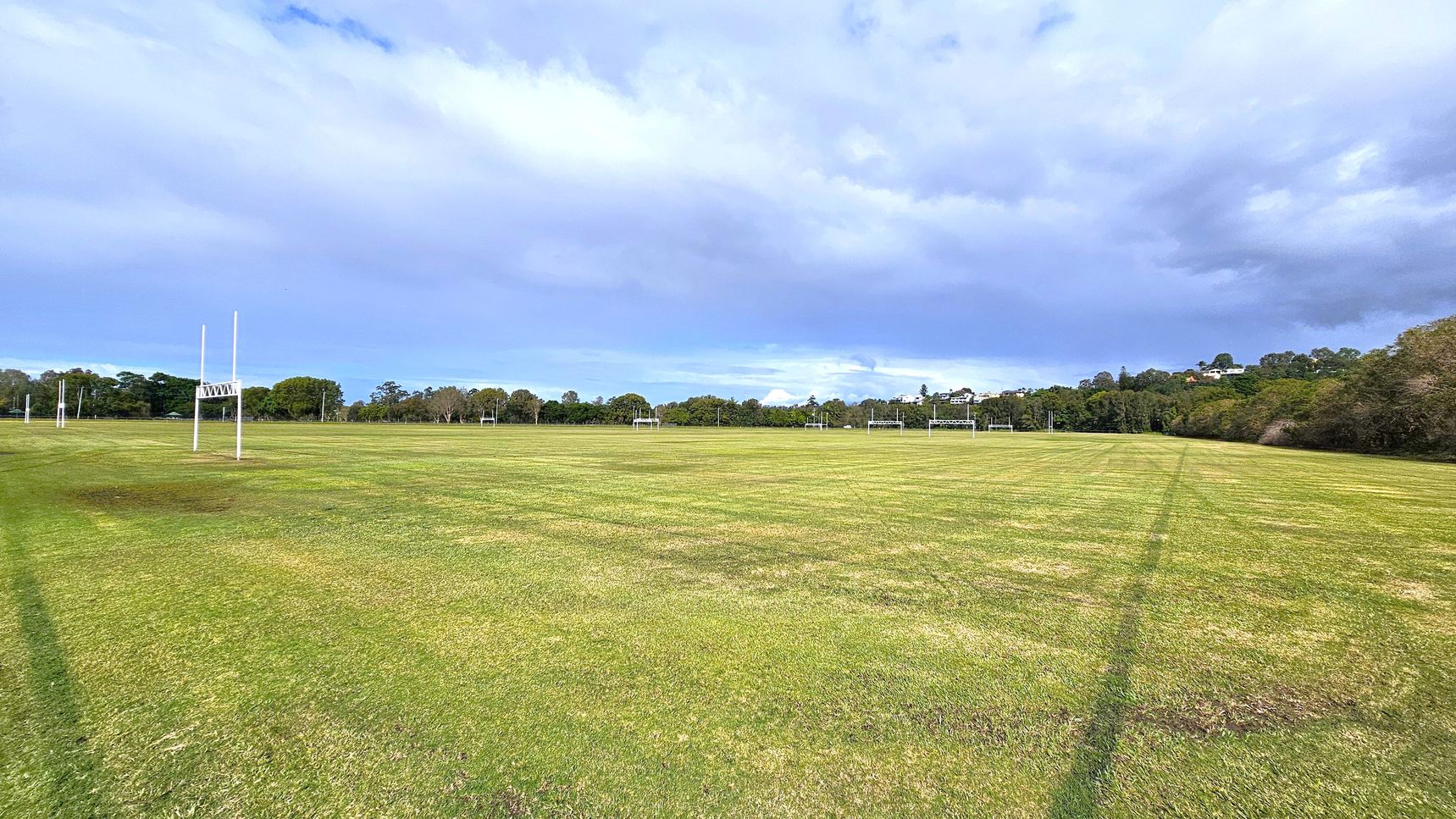 A wide, open grassy field with several rugby goalposts evenly spaced across the area. The grass is a mix of green and brown patches. In the background, there are trees and some houses visible on a slight hill. The sky is overcast with a mix of light and dark clouds.