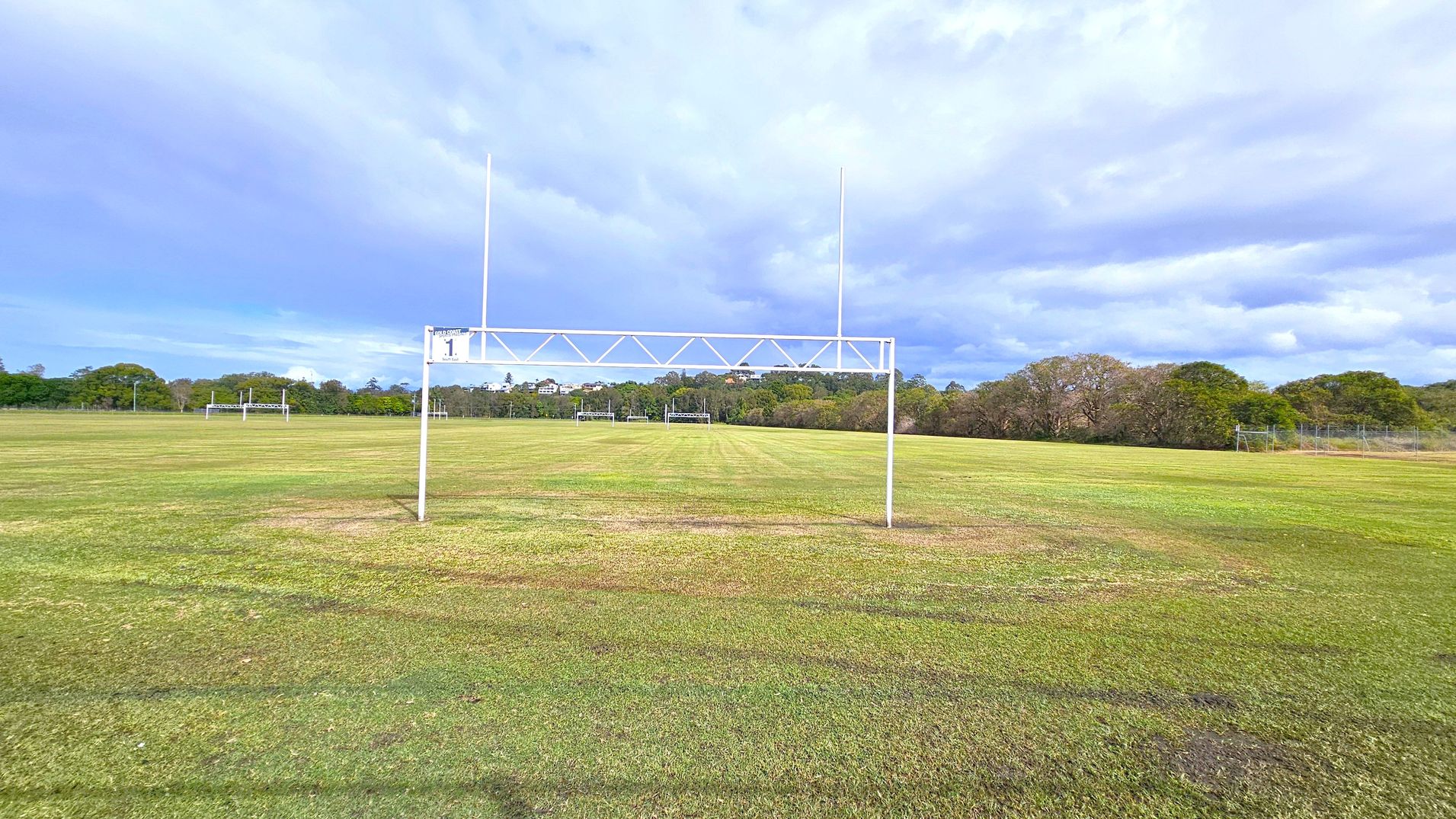 A grassy sports field with a white goalpost in the foreground, marked with the number "1". In the background, more goalposts are visible, along with a line of trees and some distant buildings. The field is expansive, with a well-maintained lawn. The sky is overcast, casting a soft light over the scene.