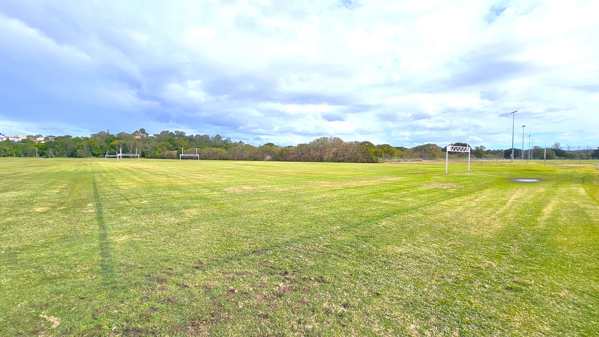 A large grassy field with rugby goalposts on the left and right sides. The grass is a mix of green and brown patches. In the background, there are trees and some distant buildings partially visible. Several tall floodlights are positioned on the right side of the field. The sky is overcast with a mix of light and dark clouds. A small puddle of water is visible on the right side of the field.