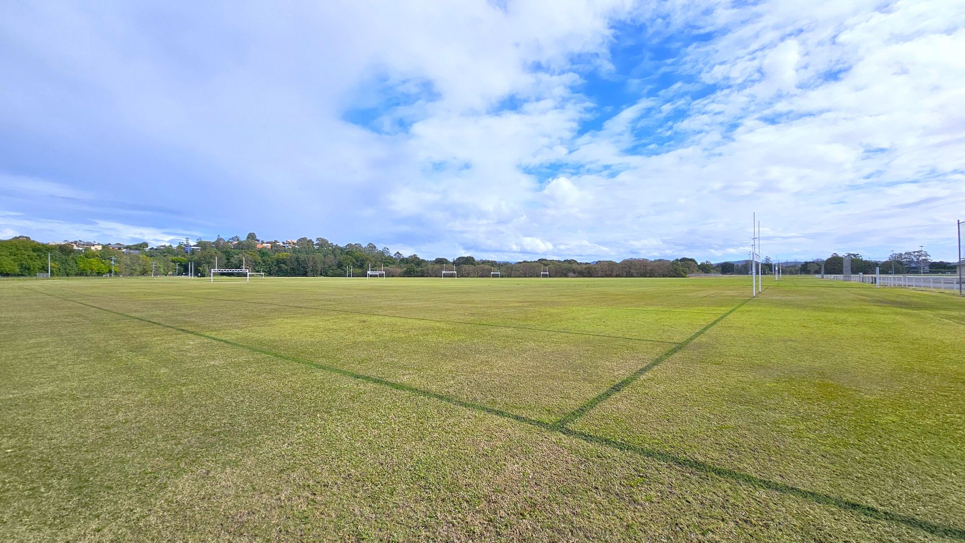 A large, open sports field with green and brown grass, marked with lines for games. Rugby posts are visible in the distance. On the left, there are trees and some buildings on a hill. The right side has a fence running along the field's edge. The background features a line of trees under a partly cloudy sky.