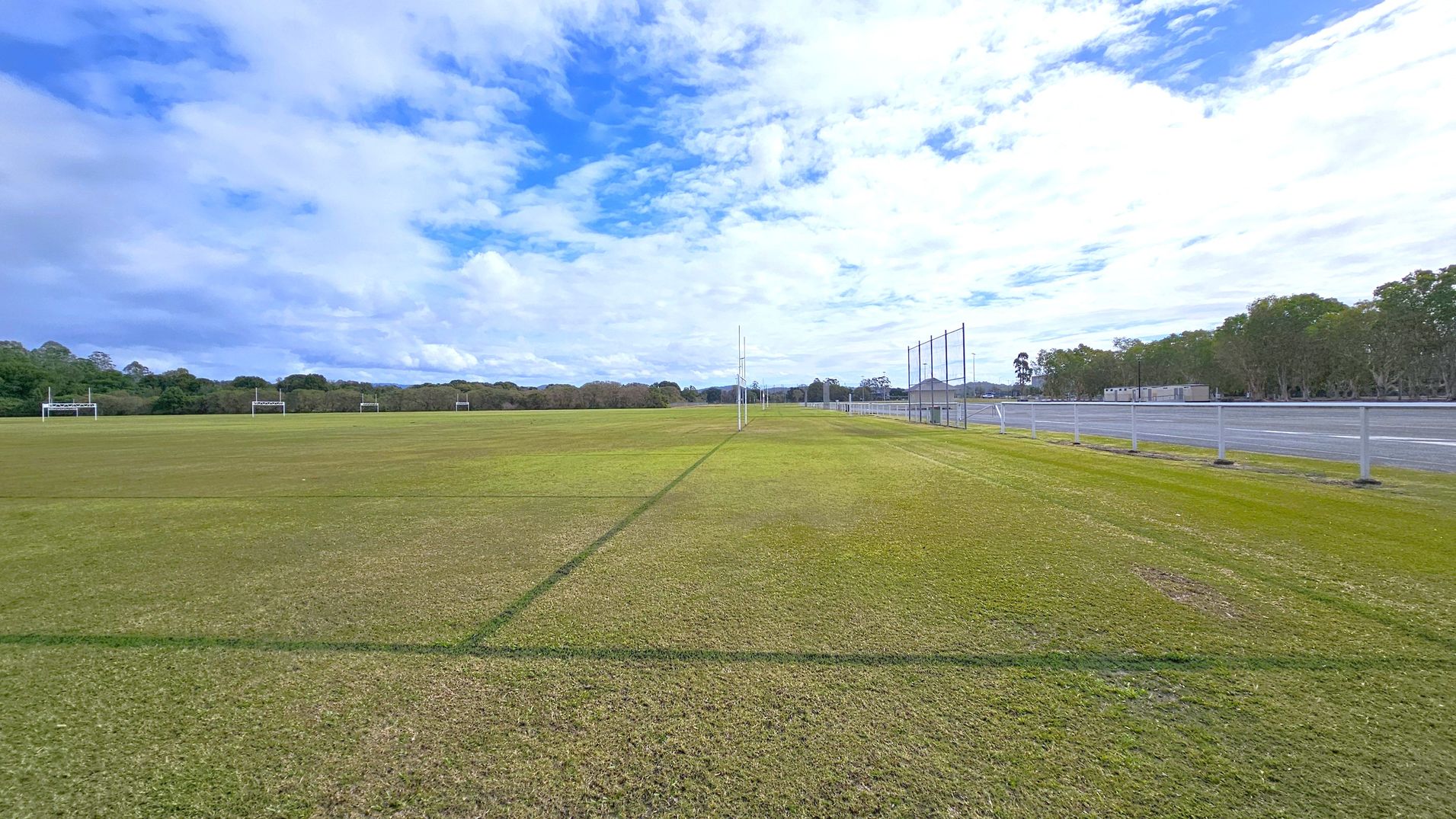 A wide, open sports field with green grass, marked with lines. In the centre, there are rugby posts. To the left, several more rugby posts are visible in the distance. On the right, a fence runs parallel to the field, with a paved area and some trees beyond it. The background features a line of trees and a few buildings. The sky is partly cloudy.