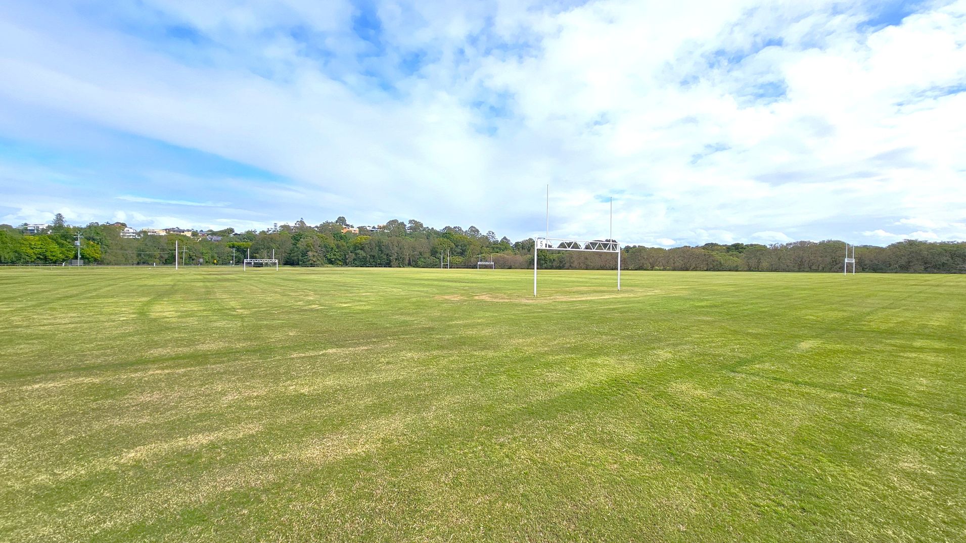 A large, open grassy field with rugby goalposts in the centre and to the right. The grass is a mix of green and brown patches. In the background, there are trees and a few buildings visible on a slight hill. The sky is mostly cloudy with some blue patches.