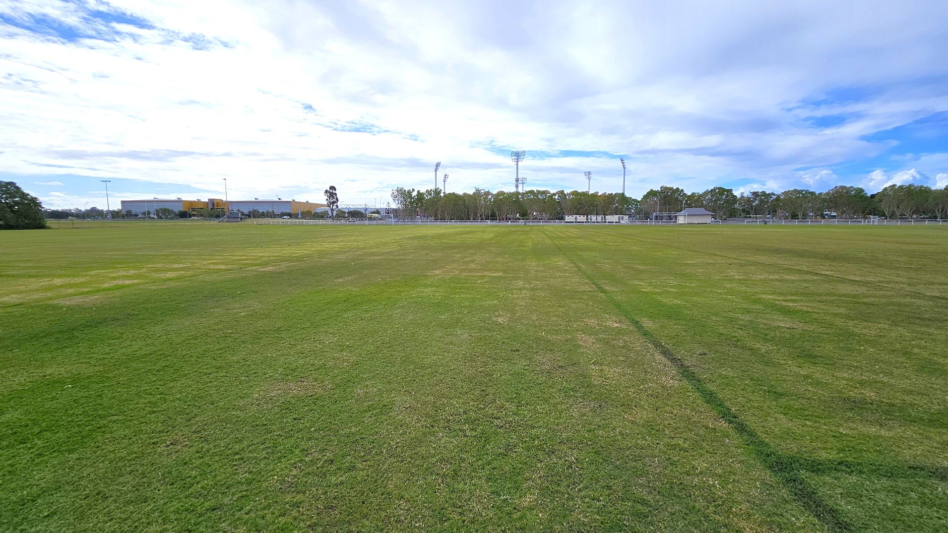 A wide, open grassy field stretches across the foreground, with patches of varying green shades. In the background, there are several tall floodlights and a line of trees. To the left, a large building with a yellow and grey facade is visible, and a few smaller structures are scattered to the right. The sky is mostly cloudy, casting a soft light over the scene.