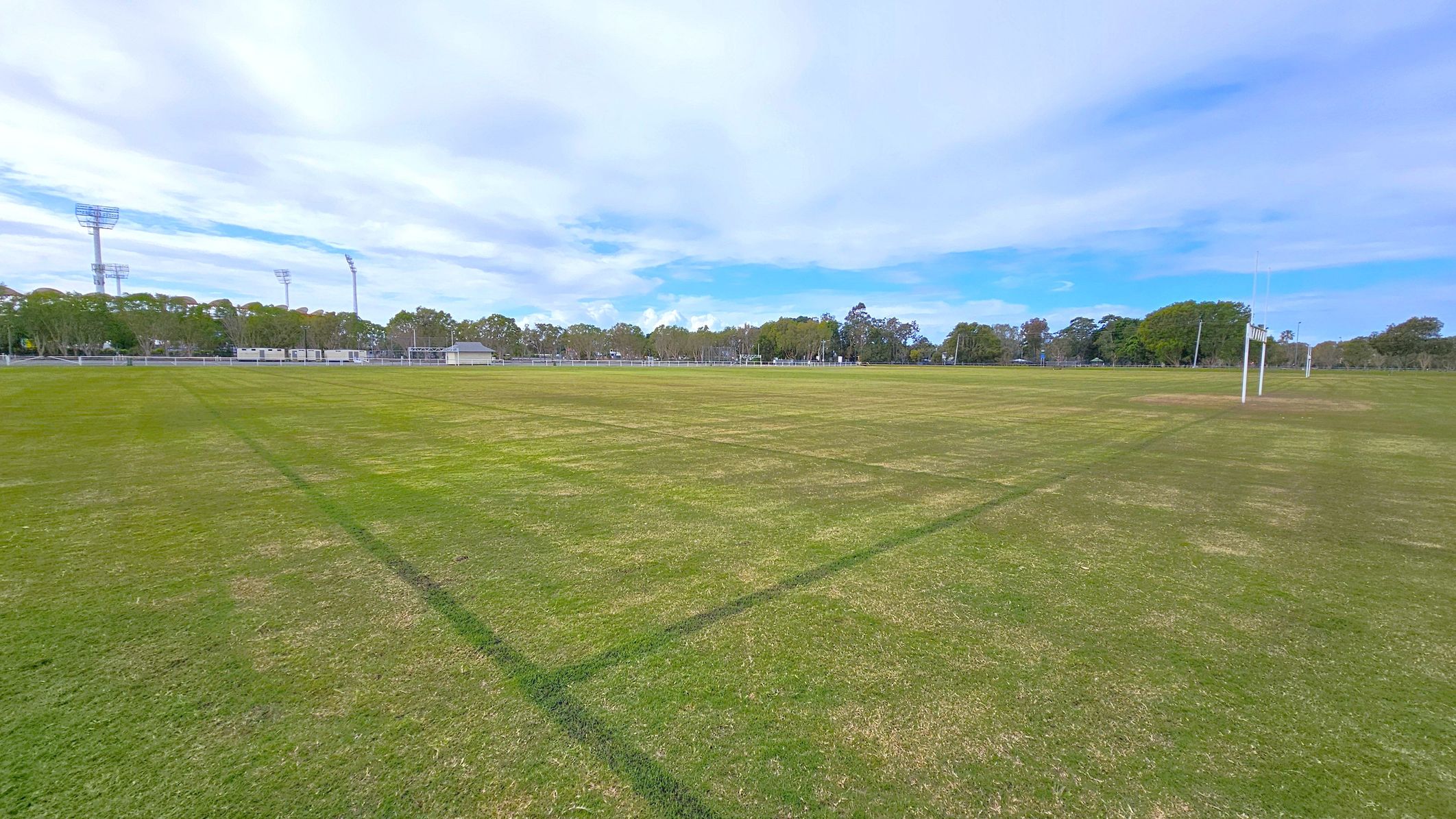 A large, open sports field with green grass, marked with faint lines. On the right, there are rugby posts. In the background, there are trees and several tall floodlights. To the left, there are some small buildings or containers near the edge of the field. The area is surrounded by more trees, creating a natural boundary.