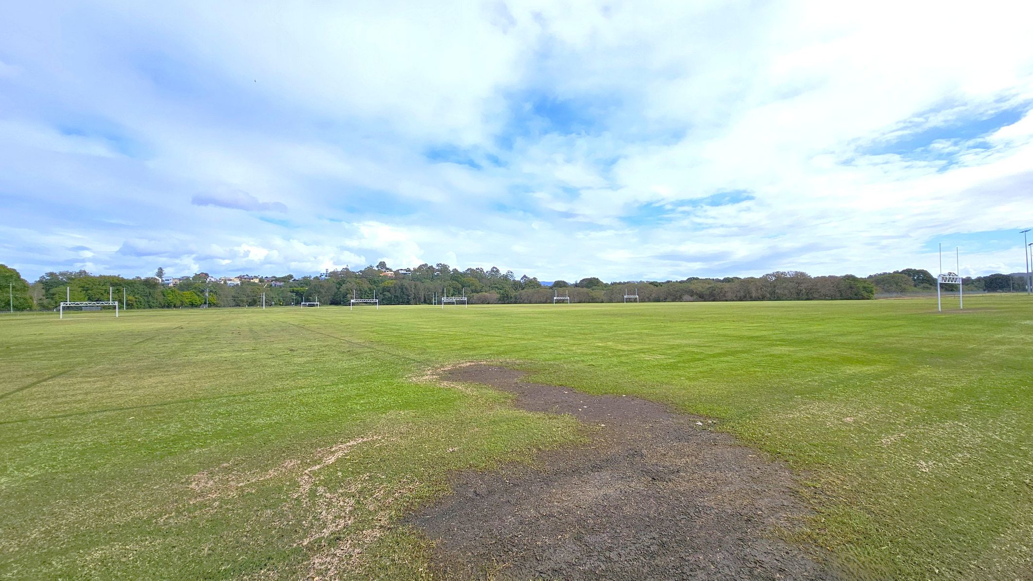 A wide, open grassy field with several rugby goalposts spread across the area. The grass is patchy in places, with some brown and bare spots visible in the foreground. In the background, there are trees and a line of houses on a slight hill. The sky is mostly cloudy with patches of blue.
