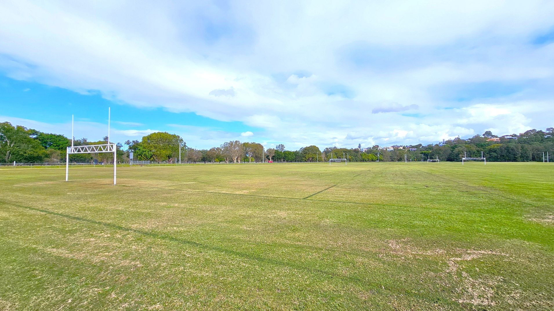 A large, open rugby field with green grass and visible pitch markings. On the left, there is a set of white rugby posts. In the background, there are more rugby posts and a line of trees with varying shades of green. The field is expansive, with a slightly uneven grass surface. The sky above is mostly cloudy with patches of blue.