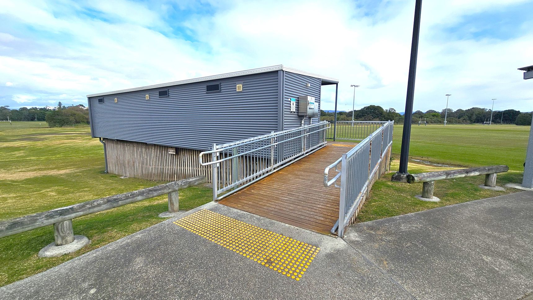 A grey corrugated metal building is elevated on wooden stilts. A wooden ramp with metal railings leads up to the building, with a yellow tactile paving strip at the base. The surrounding area is a grassy field with scattered trees in the distance. A concrete path runs alongside the ramp, and a wooden barrier is positioned near the edge of the grass. A tall black pole stands nearby, likely a light fixture.
