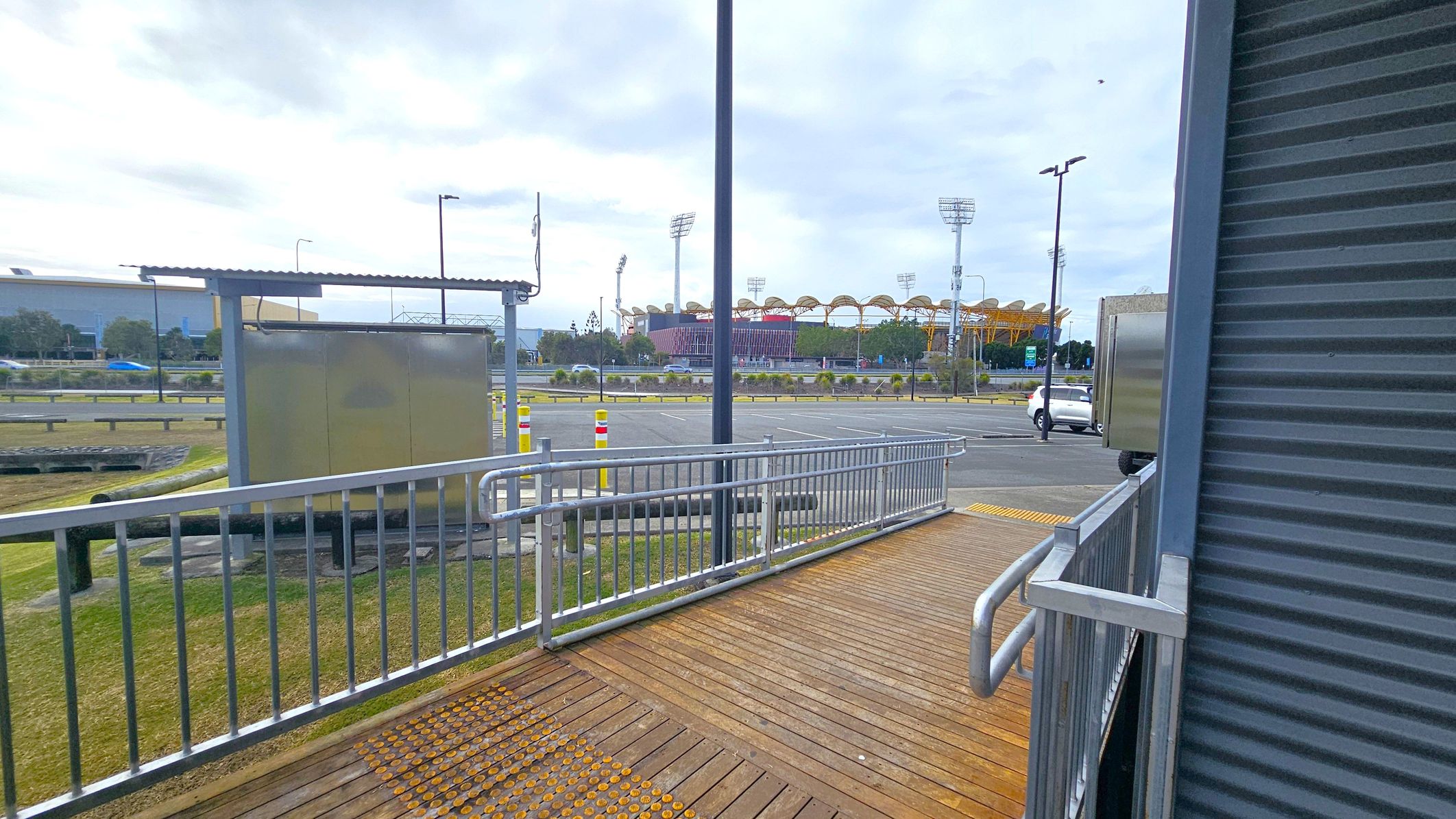 A wooden ramp with metal railings leads downwards, bordered by a corrugated metal wall on the right. In the background, a large stadium with distinctive yellow and red structures is visible. A car is parked on the road, and there are several streetlights and a small shelter with a metal roof on the left. The ground is grassy with some paved areas.