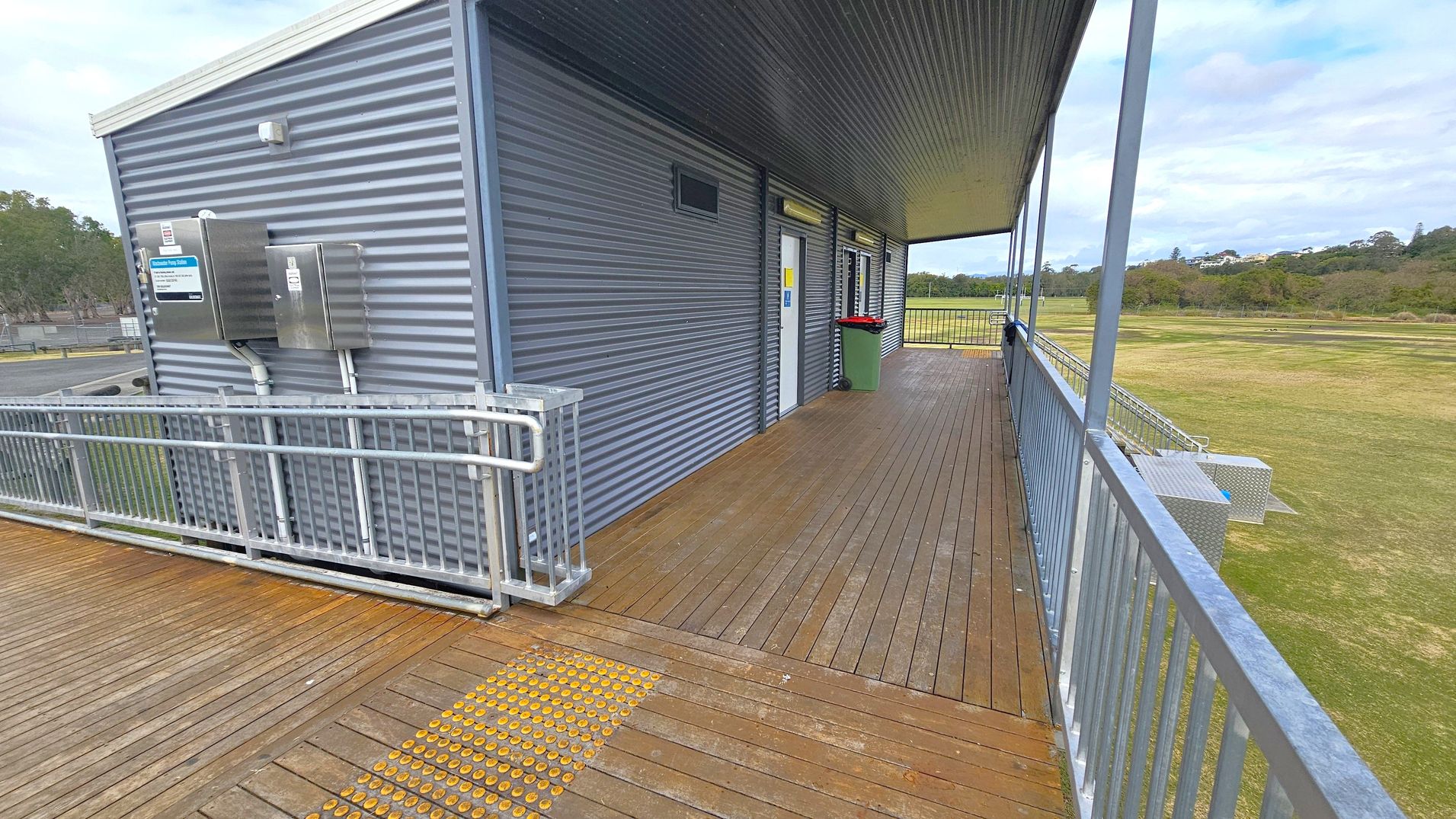 A corrugated metal building with a wooden deck and metal railing. On the left, there are two metal boxes attached to the wall. The deck has a yellow tactile paving strip. A door with a sign is visible in the centre, next to a green bin with a red lid. The right side opens to a grassy field with trees in the background. The ceiling is metal, matching the building's exterior.