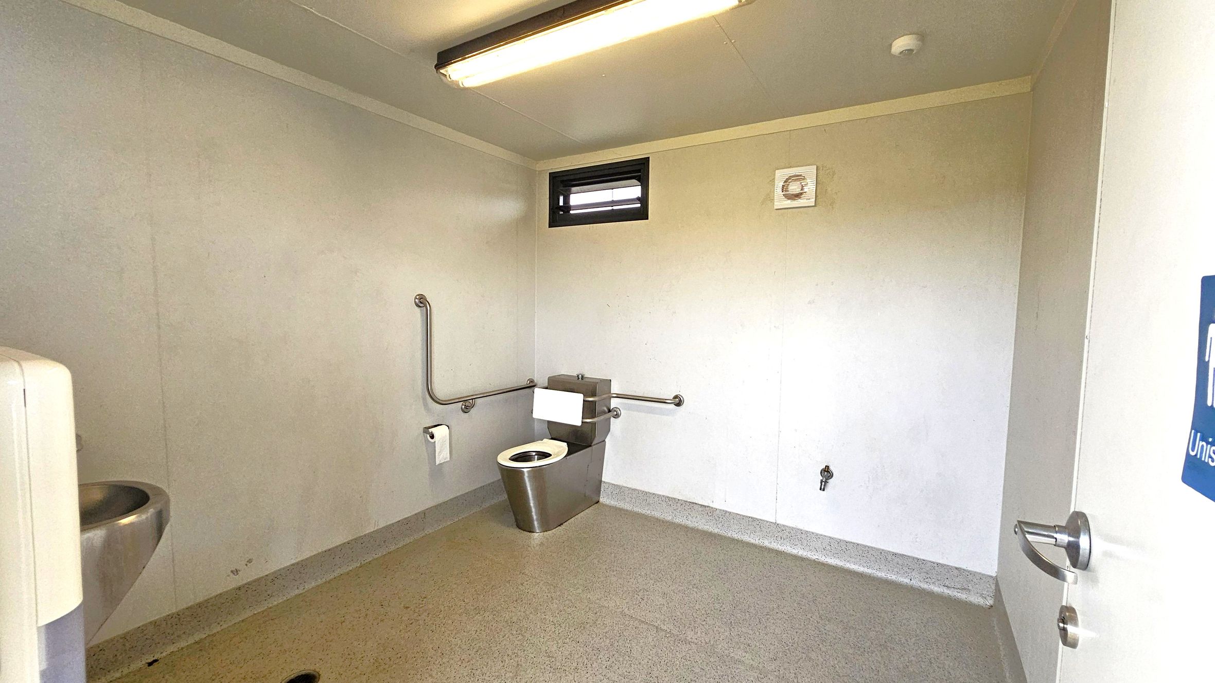 A public restroom with a stainless steel toilet in the centre, equipped with grab bars on both sides. To the left, a stainless steel sink is visible. The walls are plain and light-coloured, with a small window and a vent above the toilet. The floor is speckled grey, and the ceiling has a fluorescent light fixture. A door with a handle is partially open on the right, displaying a sign indicating a unisex toilet.