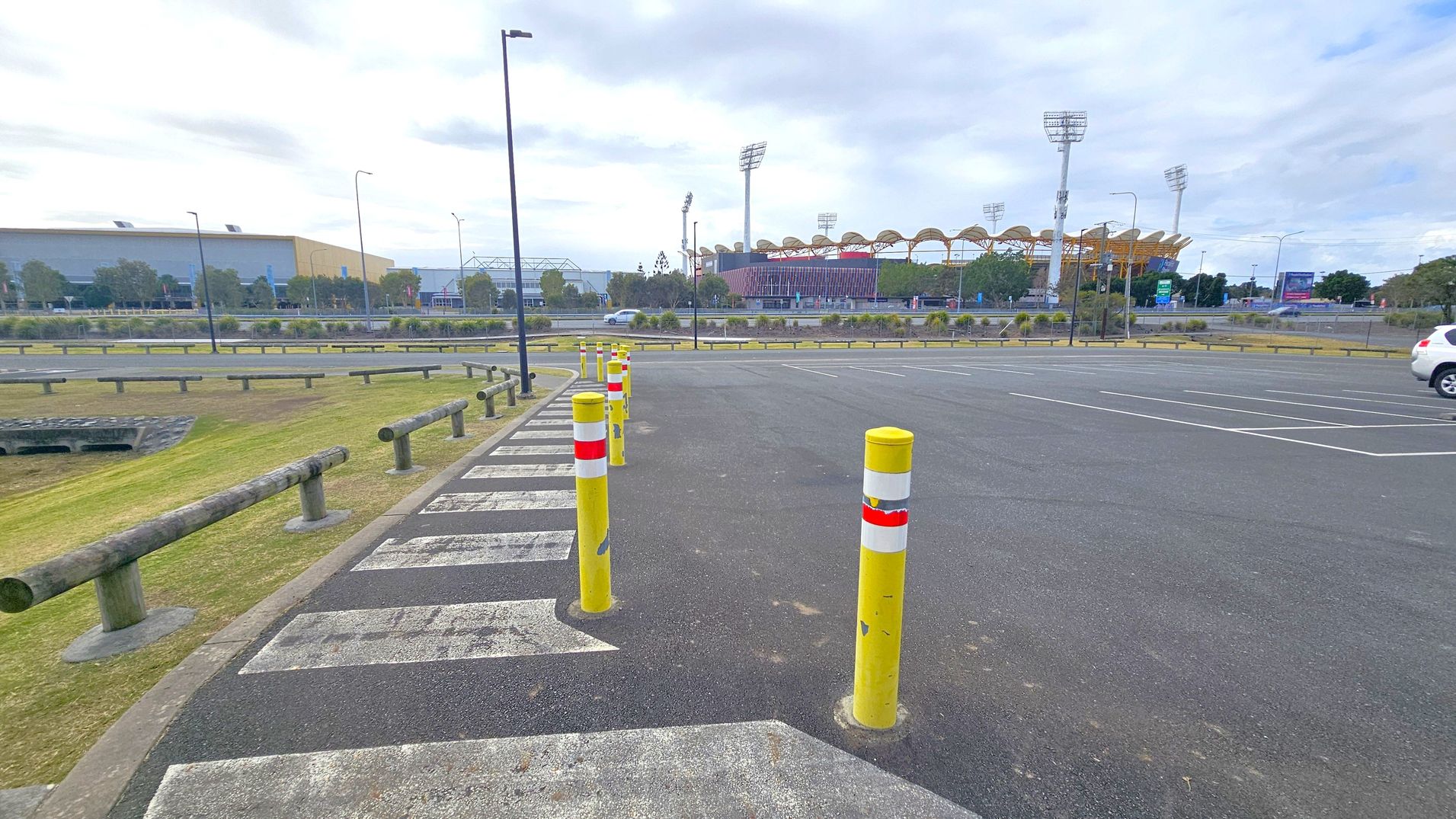 A parking area with yellow bollards lined up along a pedestrian crossing. The ground is asphalt with white painted lines. On the left, there is a grassy area with wooden barriers. In the background, a large building with a curved roof and tall floodlights is visible, likely a stadium. A few trees and shrubs are scattered around, and a white car is parked on the right side. The sky is overcast.