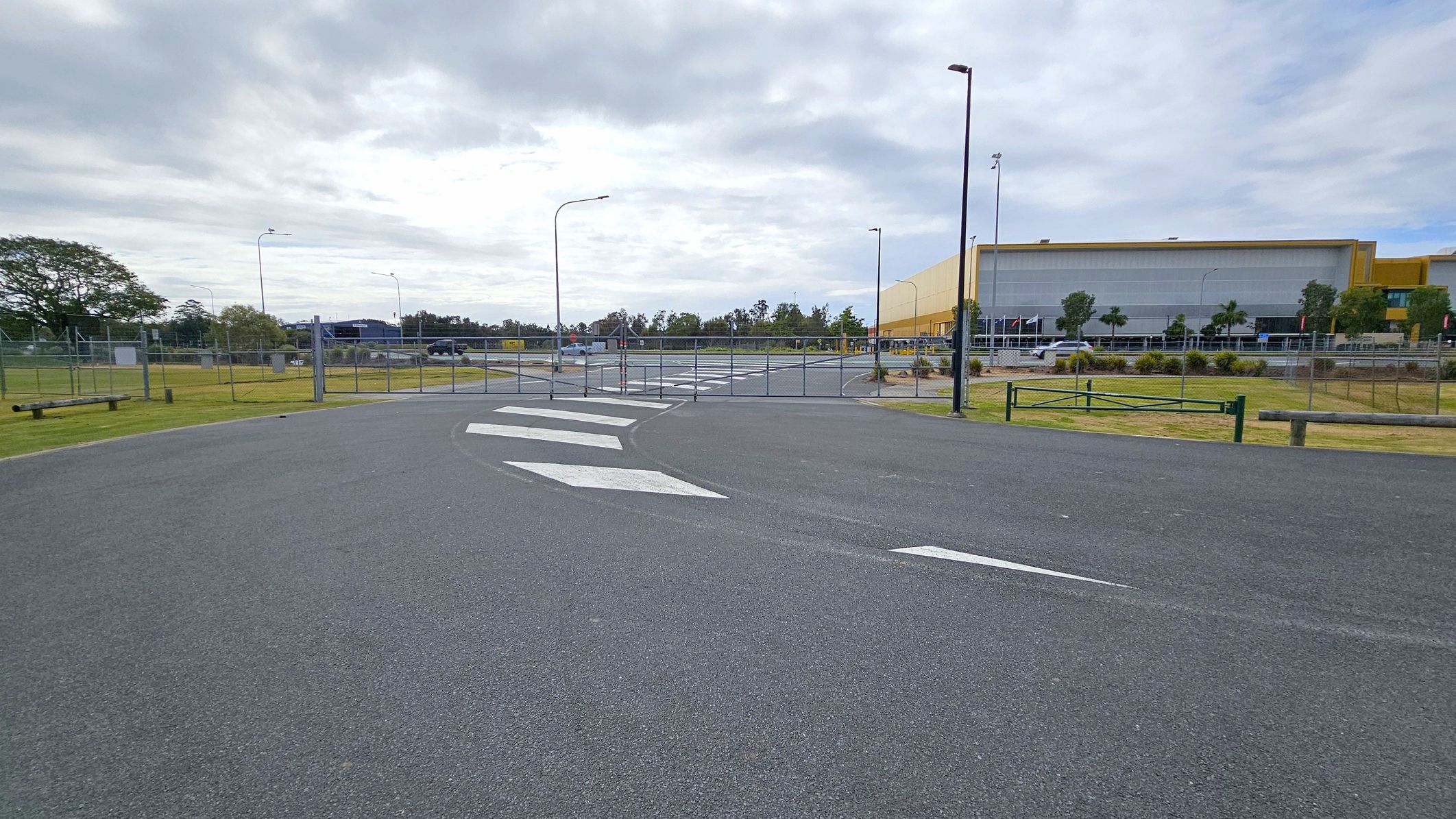 A paved road with white directional arrows leads to a closed metal gate. On the right, a large industrial building with a yellow facade is visible, surrounded by trees and a fence. On the left, there is a grassy area with a few trees and a bench. Several streetlights line the road, and a few vehicles are parked in the distance. The sky is overcast.