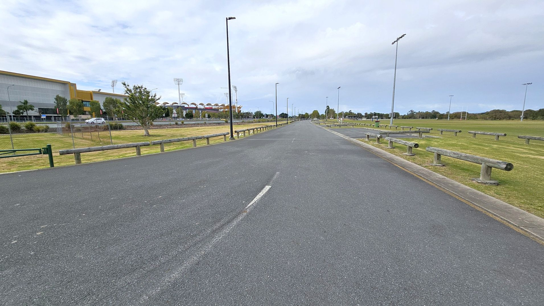 A wide, empty road stretches into the distance, flanked by wooden barriers on both sides. On the left, a large building with a modern design and a row of trees is visible, along with a parked car. On the right, there is an open grassy area with more wooden barriers and tall floodlights. The road is lined with streetlights, and the sky is overcast.
