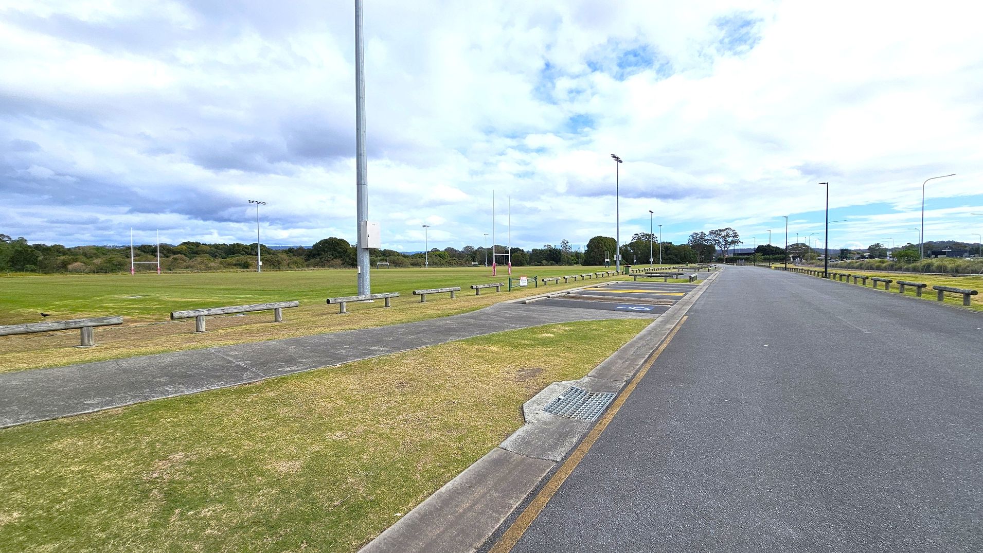 A wide open area with a rugby field on the left, featuring goalposts and surrounded by grass. A paved path runs parallel to the field, with wooden barriers separating it from the road. On the right, a long, empty asphalt road stretches into the distance, lined with streetlights and parking spaces. Trees and bushes are visible in the background, under a cloudy sky.