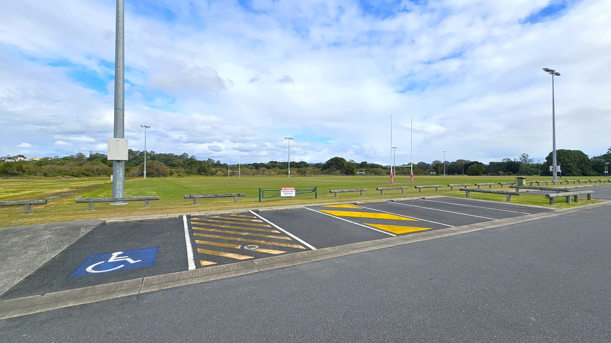 A parking area with a designated disabled parking space marked in blue on the left. Adjacent to it are spaces with yellow diagonal lines. In the background, a large grassy field with rugby posts and several tall floodlights. A sign on a green barrier reads "No Parking." The area is bordered by low wooden barriers and trees in the distance. The sky is mostly cloudy.