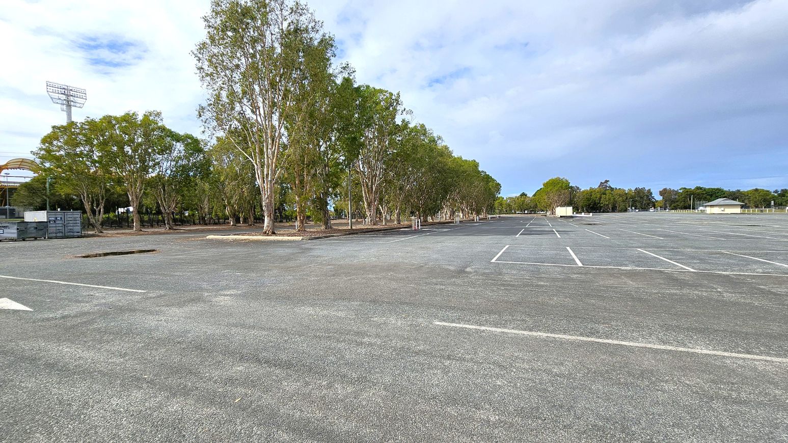 An expansive, empty car park with marked parking spaces stretches across the foreground. On the left, a row of tall trees lines the edge, with a large stadium light tower visible behind them. To the right, more trees and a few small buildings are scattered in the distance. The surface of the car park is grey and textured, with a few patches of greenery visible. The sky is overcast, casting a muted light over the scene.