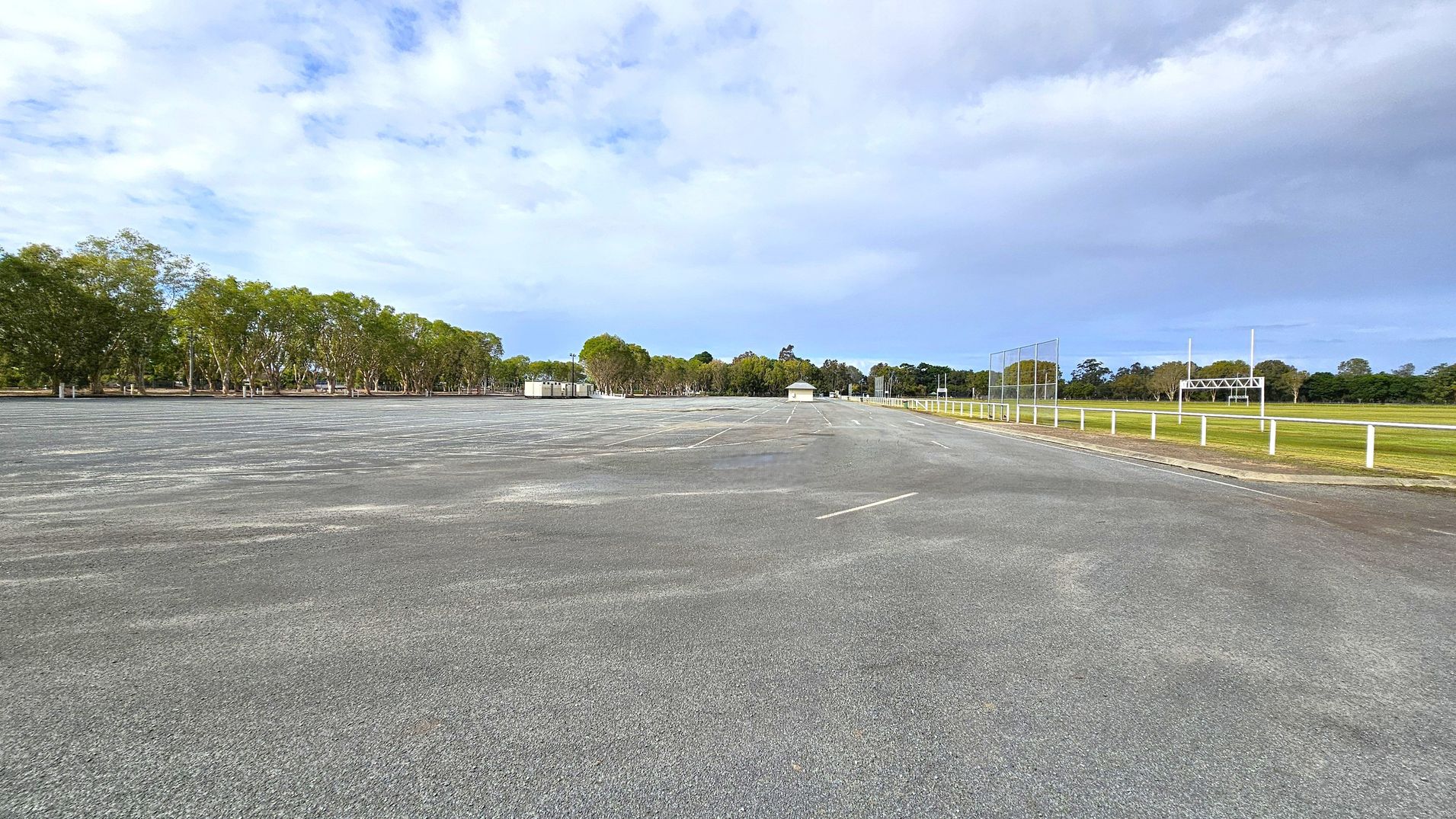 A large, empty asphalt car park stretches across the foreground. On the left, a row of trees lines the edge, providing a natural border. In the middle distance, there are small structures, possibly sheds or kiosks. On the right, a grassy field is enclosed by a white fence, with a rugby or football goalpost visible. The background features more trees, creating a green backdrop. The sky is overcast with patches of blue.