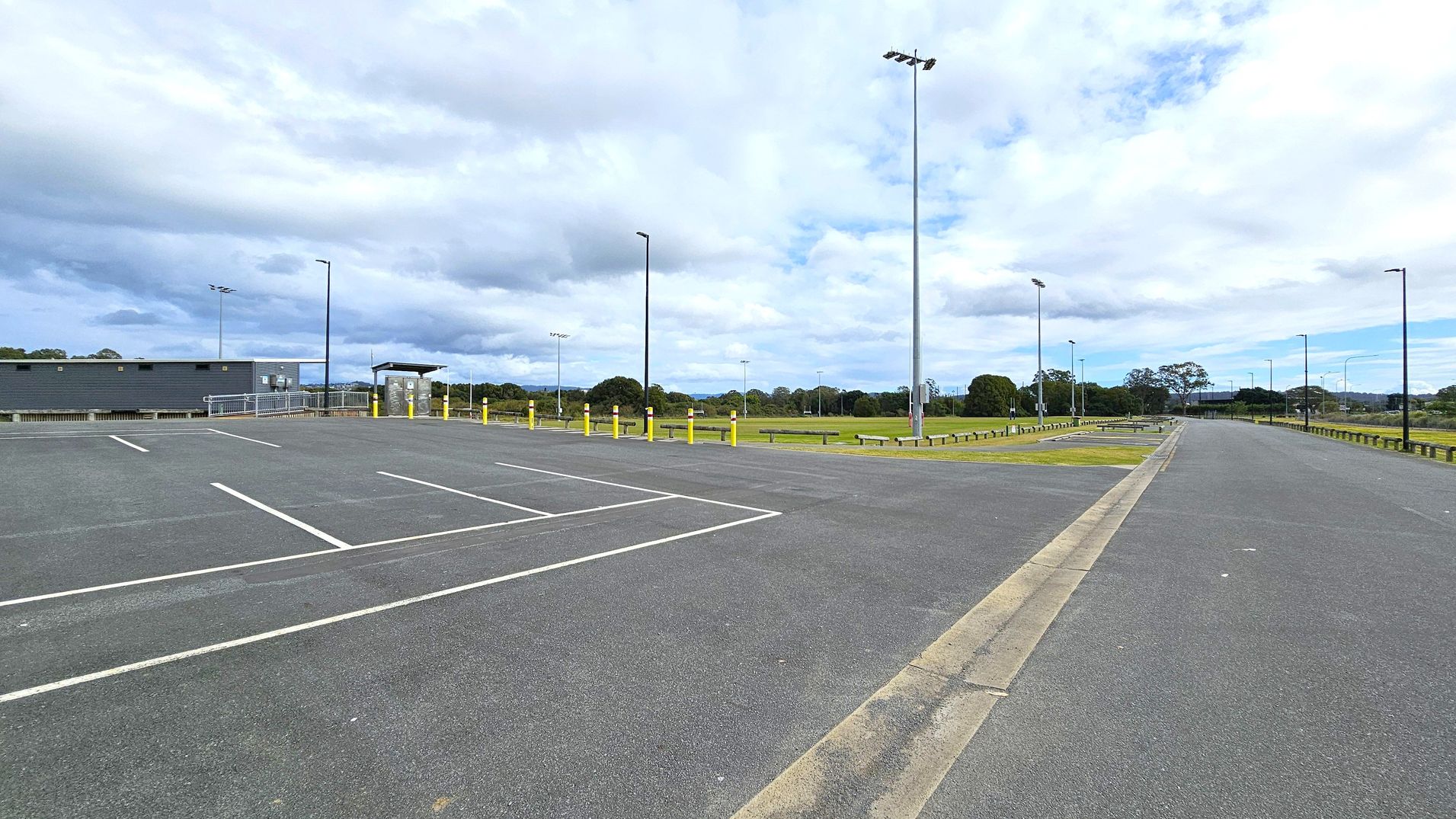 An empty car park with marked spaces on the left and a road extending into the distance on the right. A low building with a grey facade is on the far left, next to a fenced area. Several tall streetlights are scattered throughout the area. In the background, there are grassy fields and trees. The sky is overcast with clouds.