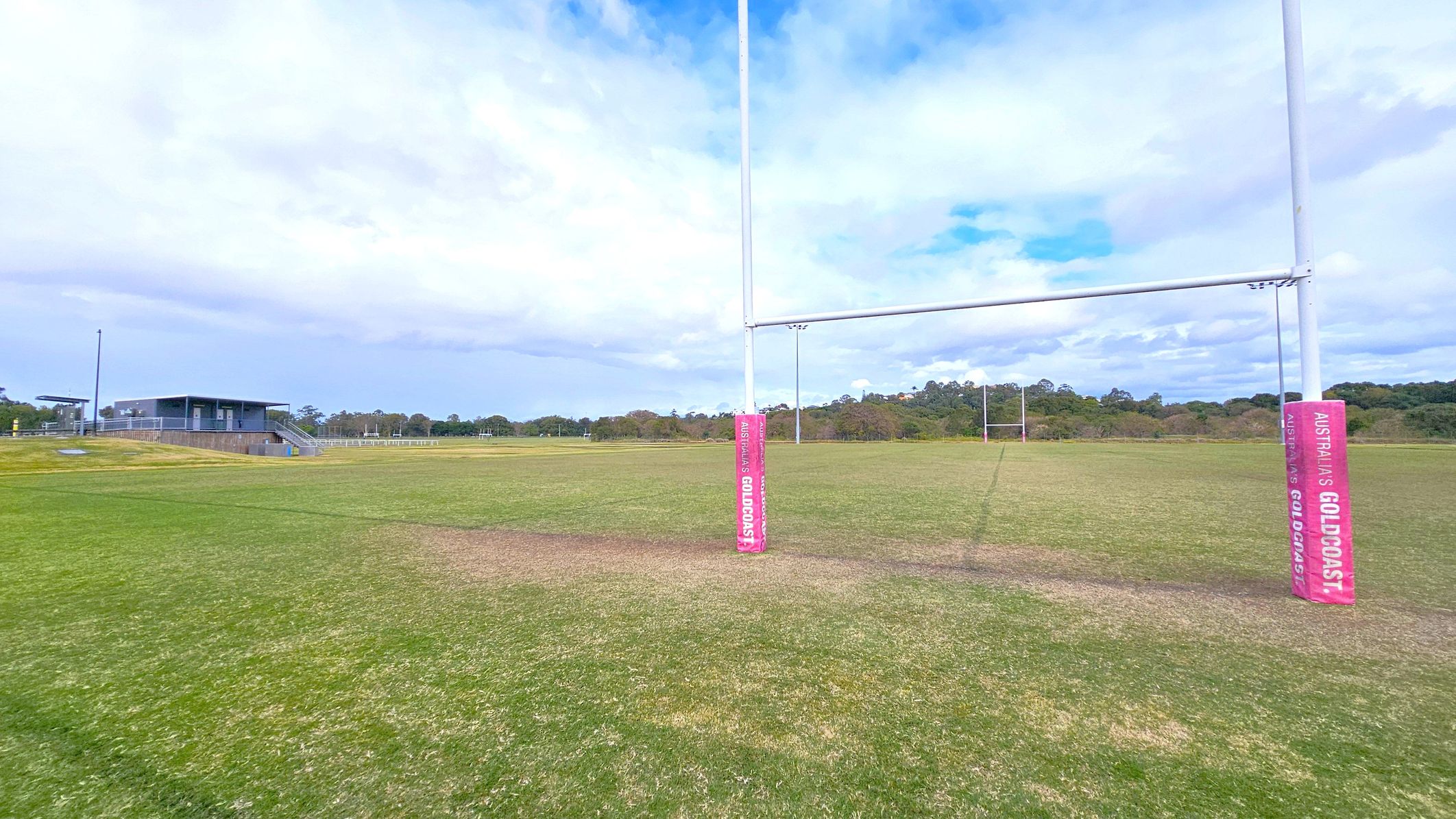 A rugby field with green grass and a set of goalposts in the centre, wrapped with pink padding labelled "Australia's Gold Coast." To the left, a small building with a staircase and a railing is visible, likely a clubhouse or viewing area. In the background, there are trees and a clear sky. The field is expansive, with another set of goalposts visible in the distance.