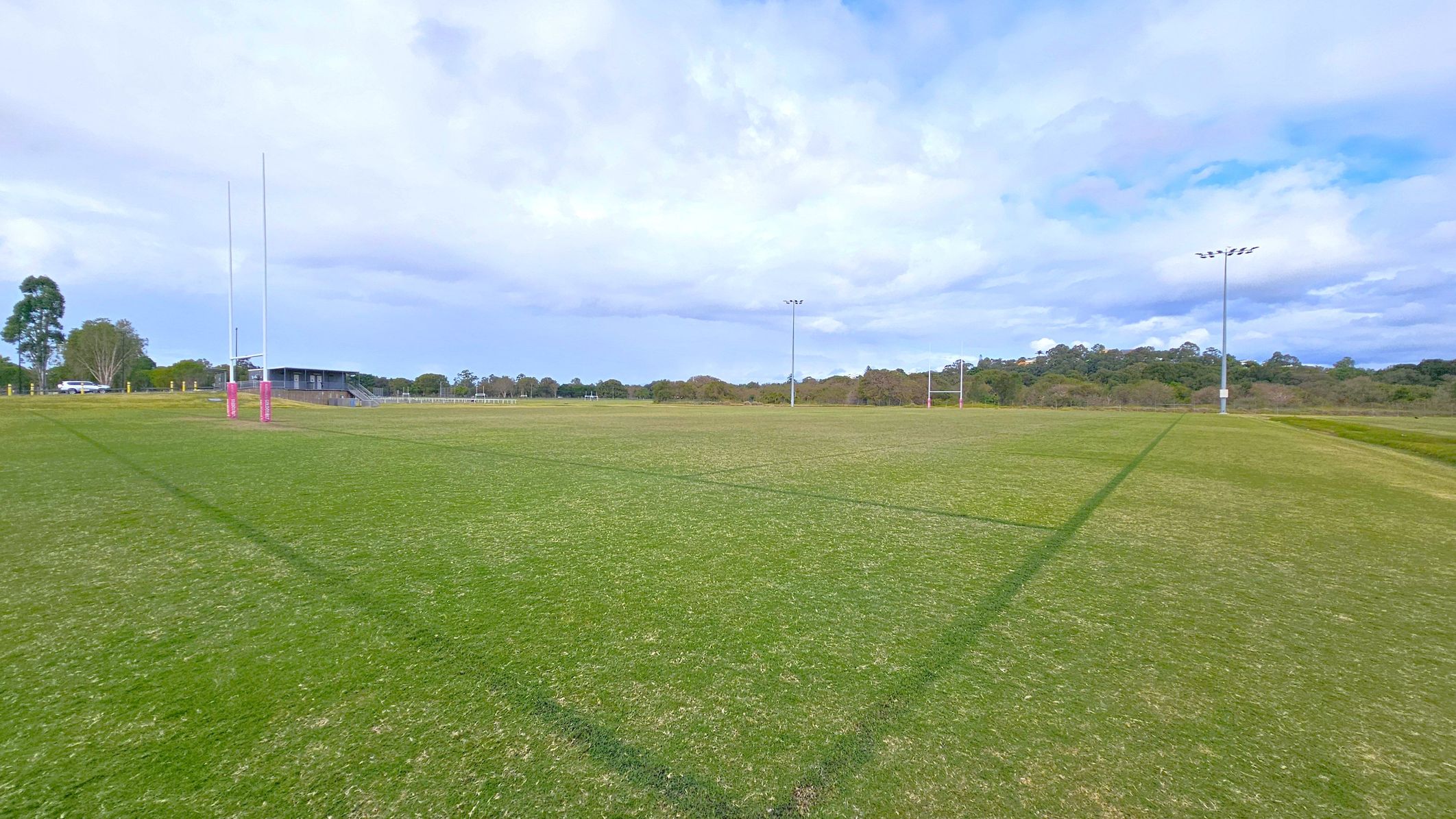 A grassy rugby field with goalposts on the left and right. The posts on the left have pink padding. A small building with a slanted roof is visible in the background on the left, near some trees and parked cars. The field is marked with dark lines, and there are tall floodlights on the right. The background features a line of trees and a slightly elevated area.