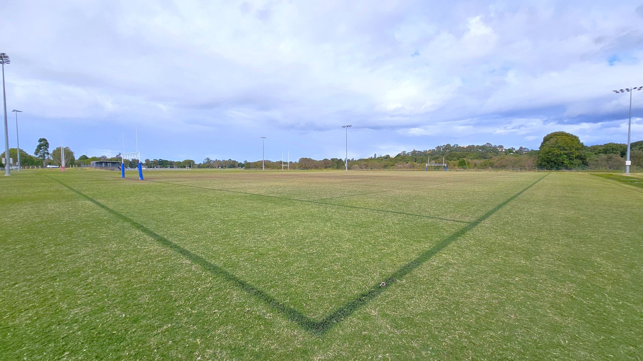A large, open sports field with green grass and marked lines. In the centre, there are rugby posts with blue padding. The field is surrounded by tall floodlights. In the background, there are trees and a small building to the left. The sky is overcast, and the field appears well-maintained.