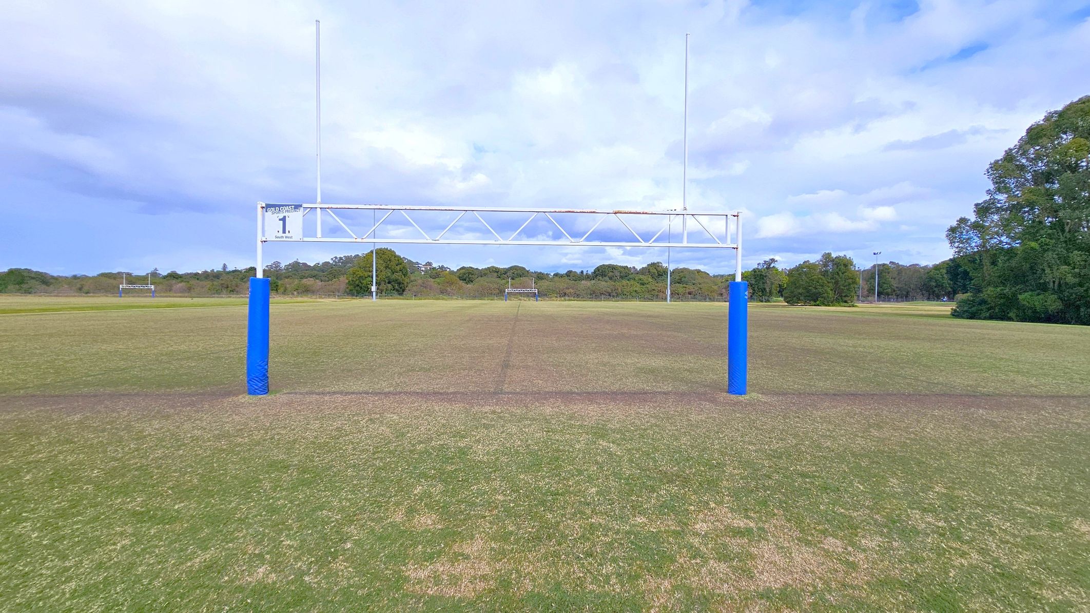 A rugby field with a goalpost in the foreground, featuring blue padding on the posts. The grass is patchy with areas of brown and green. In the background, there are more goalposts and a line of trees. The sky is overcast with clouds.