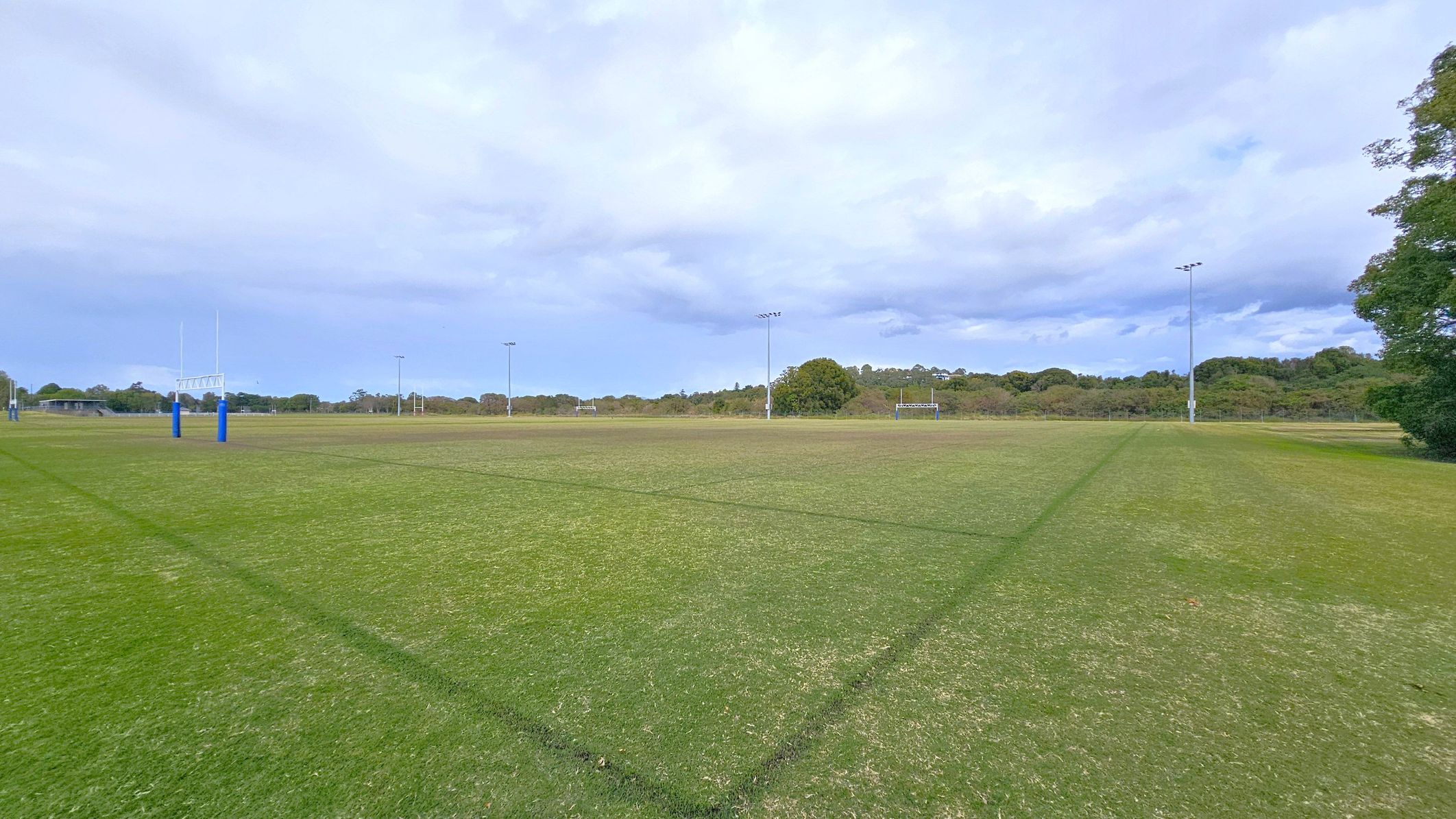 A grassy sports field with rugby posts on the left, marked by blue padding. The field is lined with green markings. In the background, there are several tall floodlights and a line of trees. A small building is visible to the far left. The field is expansive, with a mix of green and brown patches on the grass.
