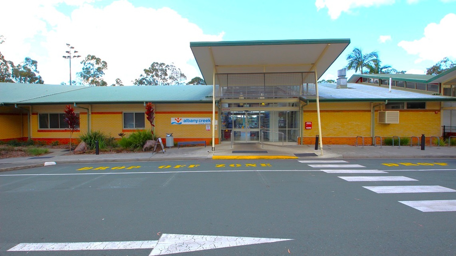 Main entrance of Albany Creek Leisure Centre. Outside the main entrance, there's visible yellow and white road marking paint. The building itself is a small, brown brick structure with a sloping corrugated iron roof. You can spot hand railings leading to a glass door at the entrance. On the left side of the entrance area, there is signage indicating "Albany Creek."