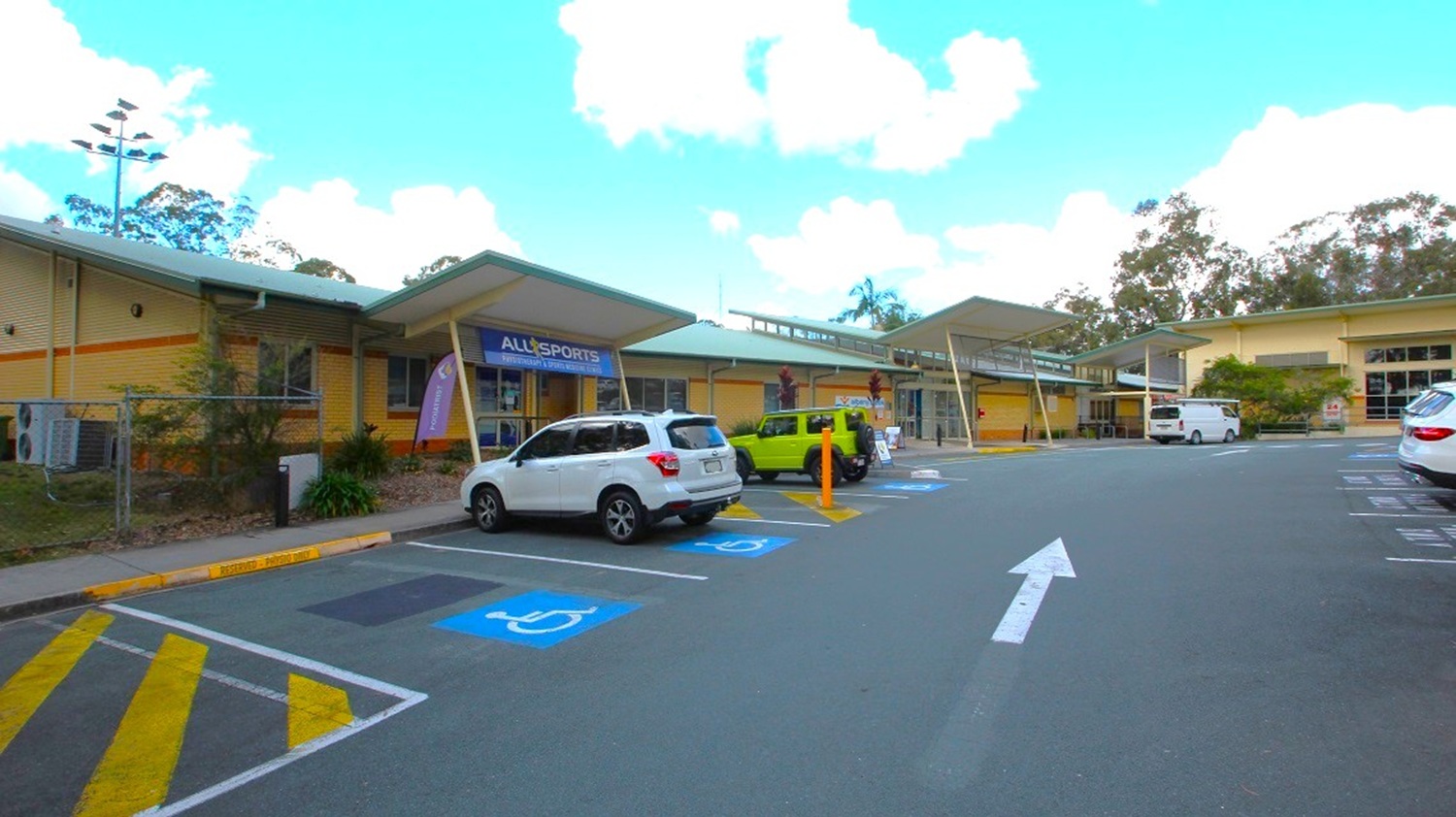 View of one of the accessible car parks of the open-air aquatic centre. Many cars are visibly parking in the parking area, with gum and pine trees in view at the rear. Two vehicles are using the designated parking spaces for people with disabilities, leaving just one of those spaces unoccupied. Additionally, there is a white arrow marking the road between these accessible parking spots.