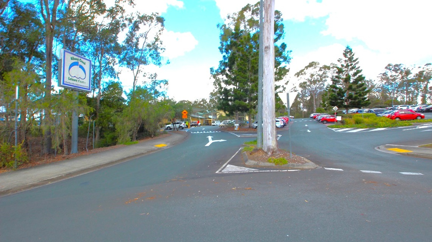 Pathway toward Albany Creek’s parking spaces. The space is expansive and well-arranged, featuring a variety of road markings with different symbols like arrows and dashed lines. Below the gum trees, there's a prominent billboard sign displaying "Albany Creek." Numerous cars are parked, and these parking areas are shaded by large pine and gum trees. Additionally, signage is strategically positioned at several intersections along the roads.