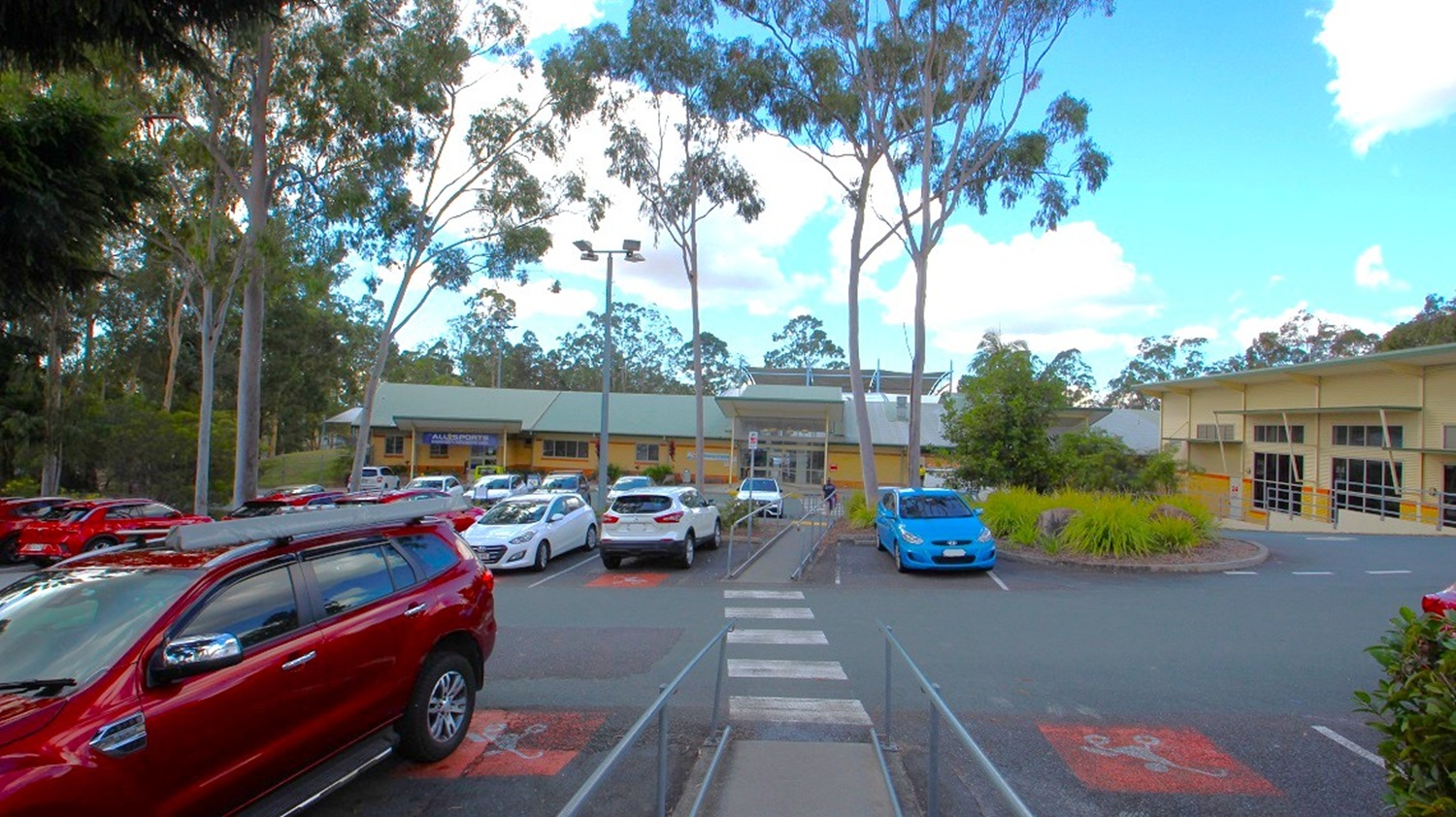 A sidewalk in the accessible parking space of Albany Creek Leisure Centre. The parking lot is enveloped by towering gum trees, providing ample shade, and it accommodates a substantial number of parked cars. Safety hand railings are strategically positioned along the parking lot's sidewalks for security. Inside the parking area, there is a massive post equipped with commercial lighting that stands prominently.